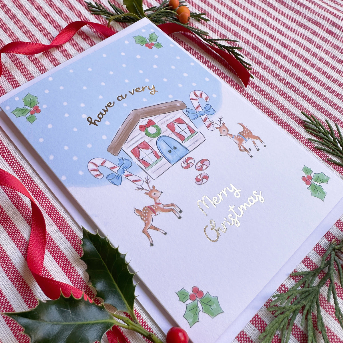 A Christmas greeting card with a whimsical house illustration, reindeer, candy canes, holly, and gold 'have a very Merry Christmas' script, styled on a red-and-white striped fabric with pine branches and a red ribbon.