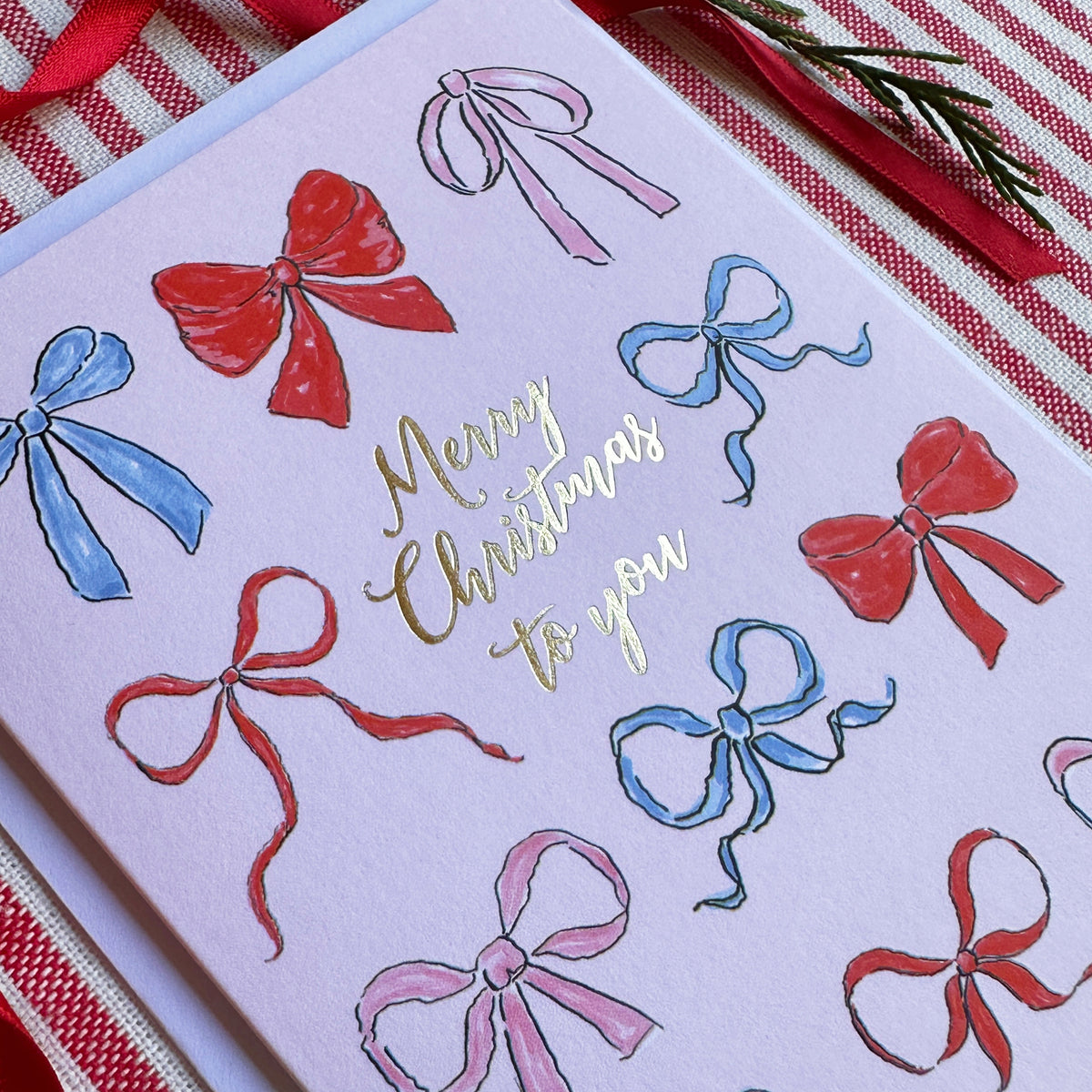 close up of a a christmas card with a soft pink background with several illustrated bows and captioned Merry Christmas to You. Background of image is red ribbon, and foliage on a red and white striped backgroumd