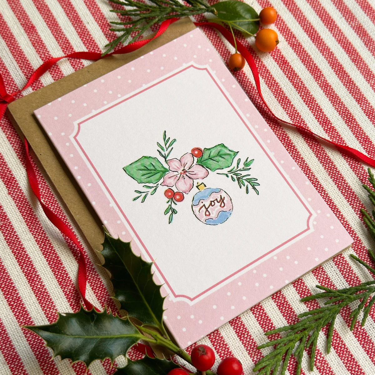 A pink Christmas greeting card with a scalloped border and a floral ornament illustration, shown with a brown envelope on a red-and-white striped fabric background with holly and ribbon.