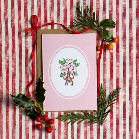 A pink greeting card with a white oval center featuring a bouquet illustration, styled on a red-and-white striped fabric with holly, pine branches, and red ribbon.