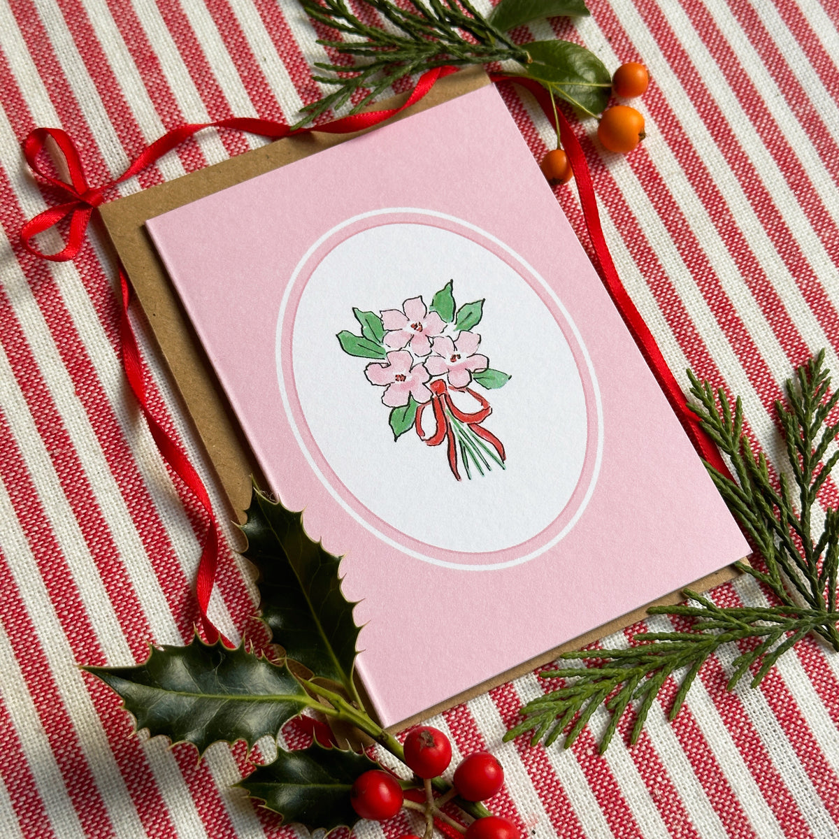 A pink greeting card with a white oval center featuring a bouquet illustration, placed on a red-and-white striped fabric with green pine branches, holly leaves, and red berries. A brown envelope is visible behind the card.