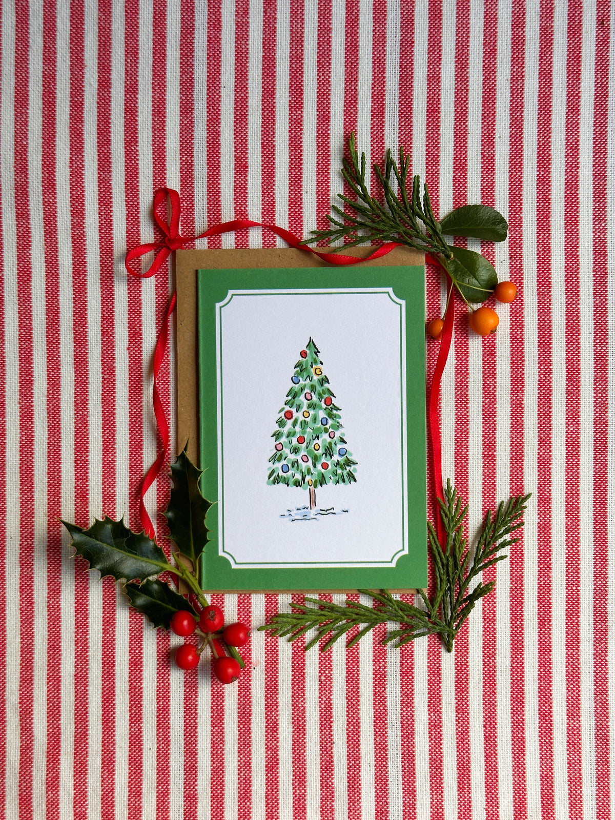 A mini Christmas greeting card with a green border and an illustration of a decorated tree, styled on a red-and-white striped fabric with pine branches, holly leaves, and red ribbon.