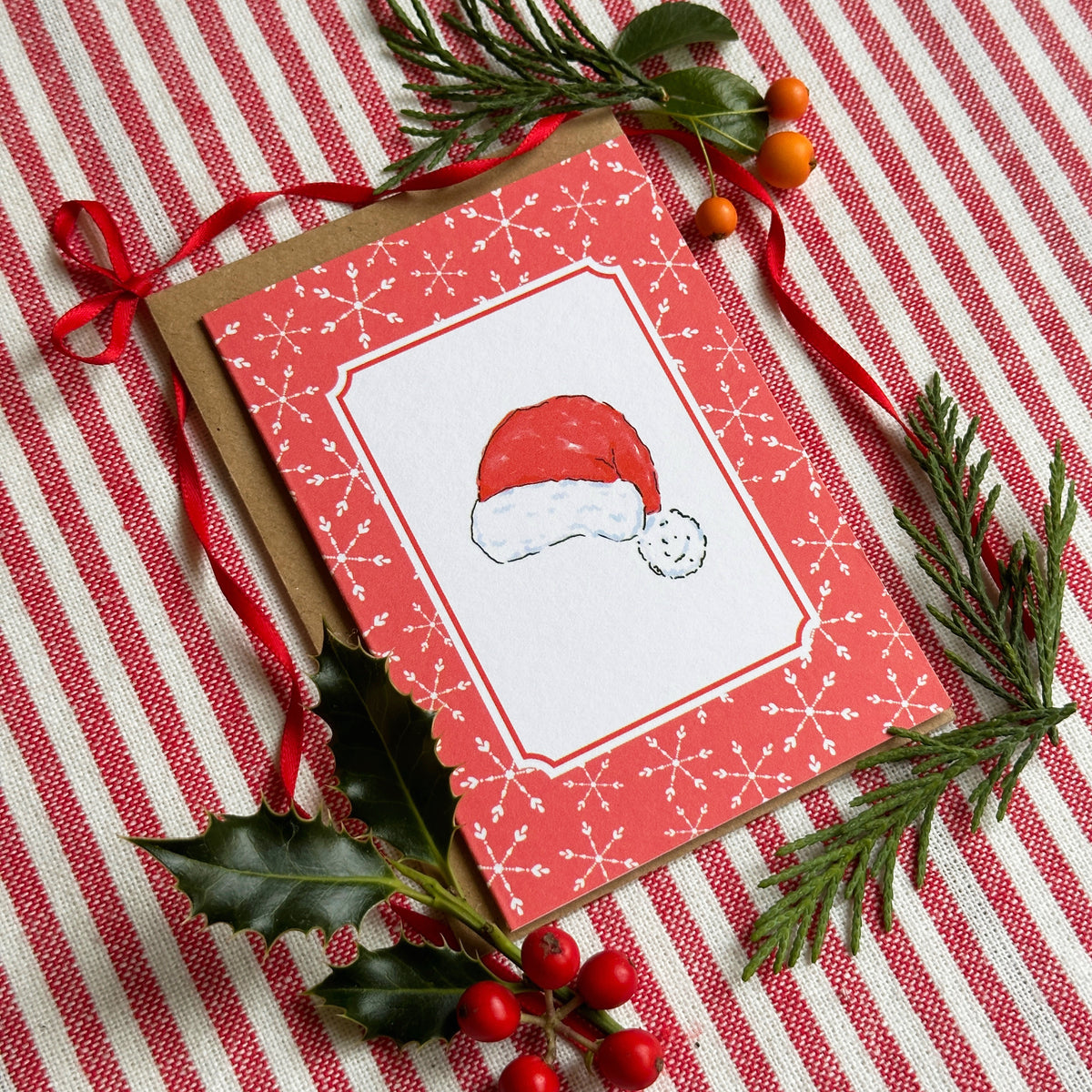 A Christmas greeting card with a red border, white center, and a Santa hat illustration, styled on a red-and-white striped fabric with greenery and berries.