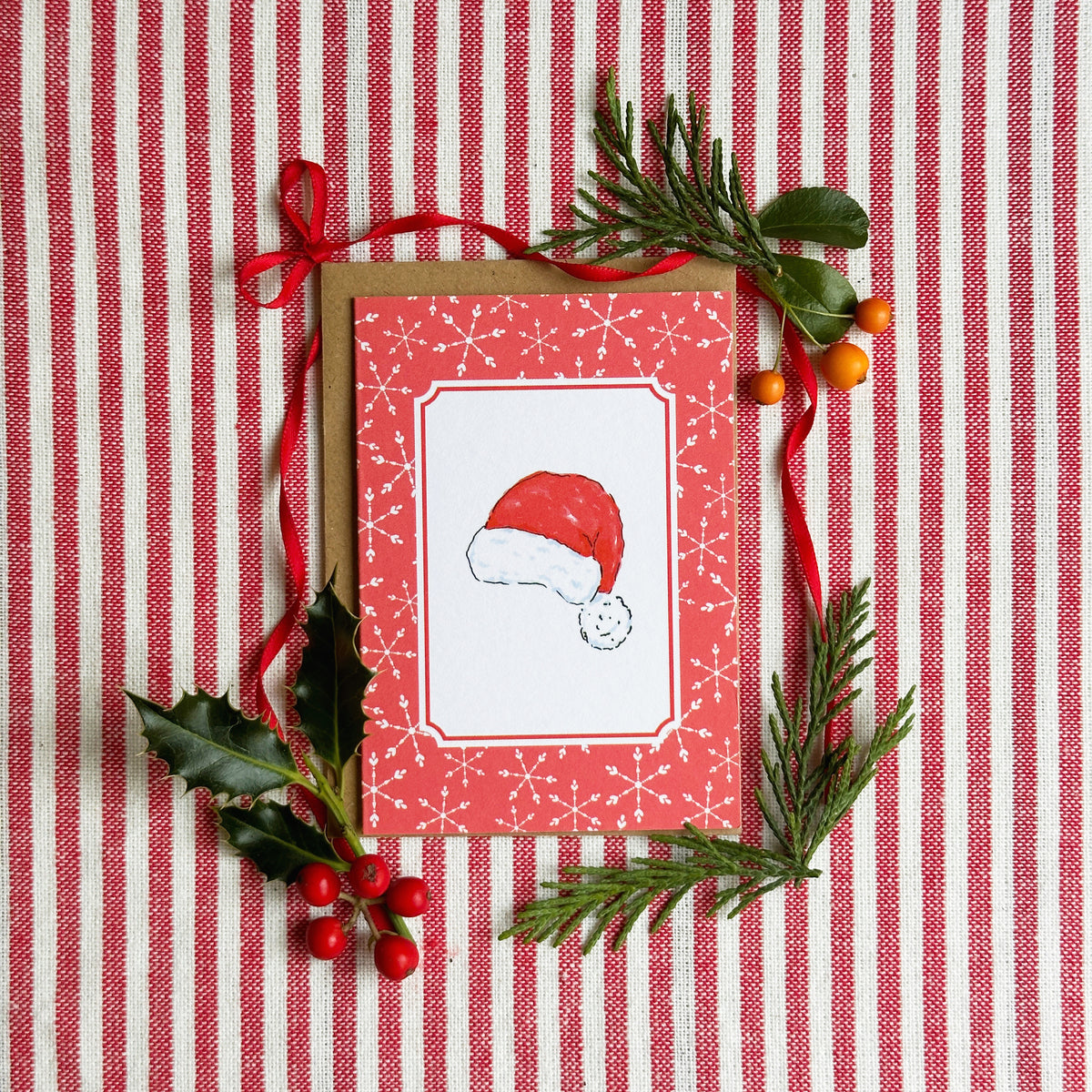 A mini A7 Christmas greeting card with a red border and a Santa hat illustration, placed on a red-and-white striped fabric background and styled with green pine branches, holly leaves, and red berries.