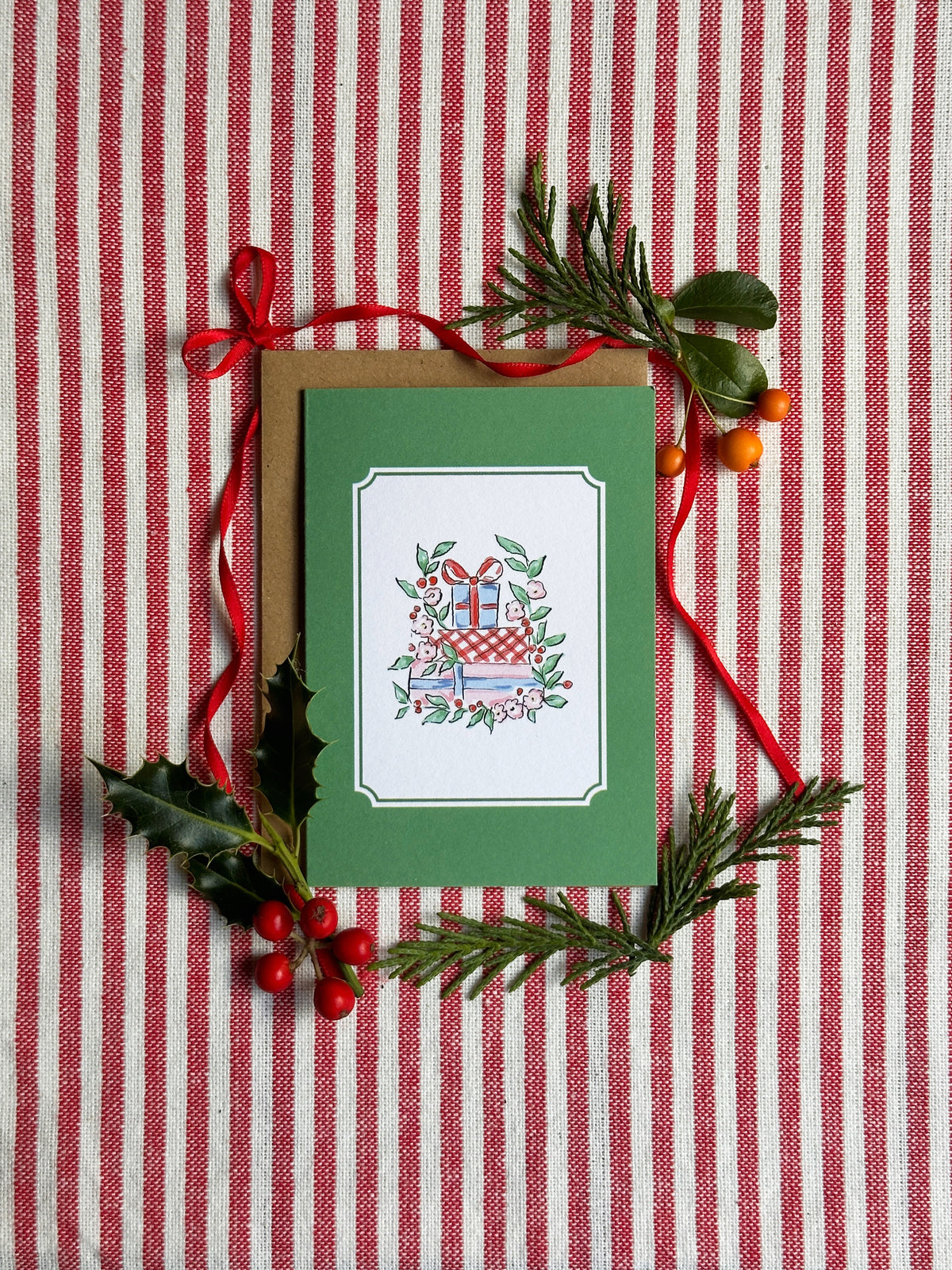 A green Christmas greeting card with a white border and an illustration of wrapped gifts, placed on a brown kraft envelope and styled with red ribbon, holly leaves, pine branches, and red berries on a red-and-white striped fabric background.