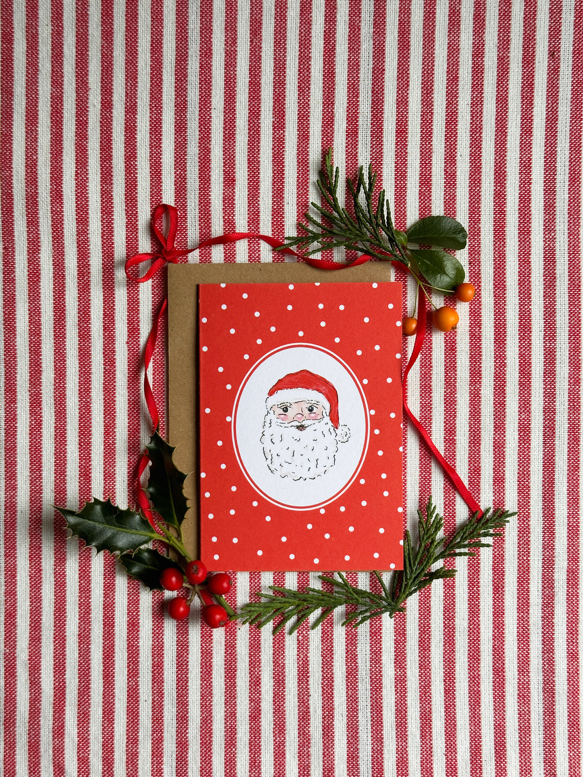 A red Christmas greeting card featuring an illustration of Santa Claus inside a white oval, layered over a brown envelope. The card is decorated with a red ribbon, green pine branches, holly leaves, and small red berries, all arranged on a red and white striped fabric background.