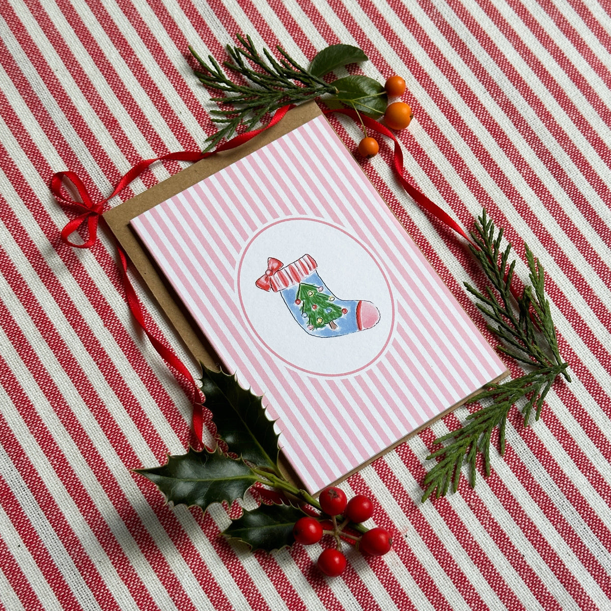 A mini Christmas greeting card with pink and white vertical stripes and a circular illustration of a blue stocking with a green tree, styled on a red and white striped fabric background with pine branches, holly, and red berries.