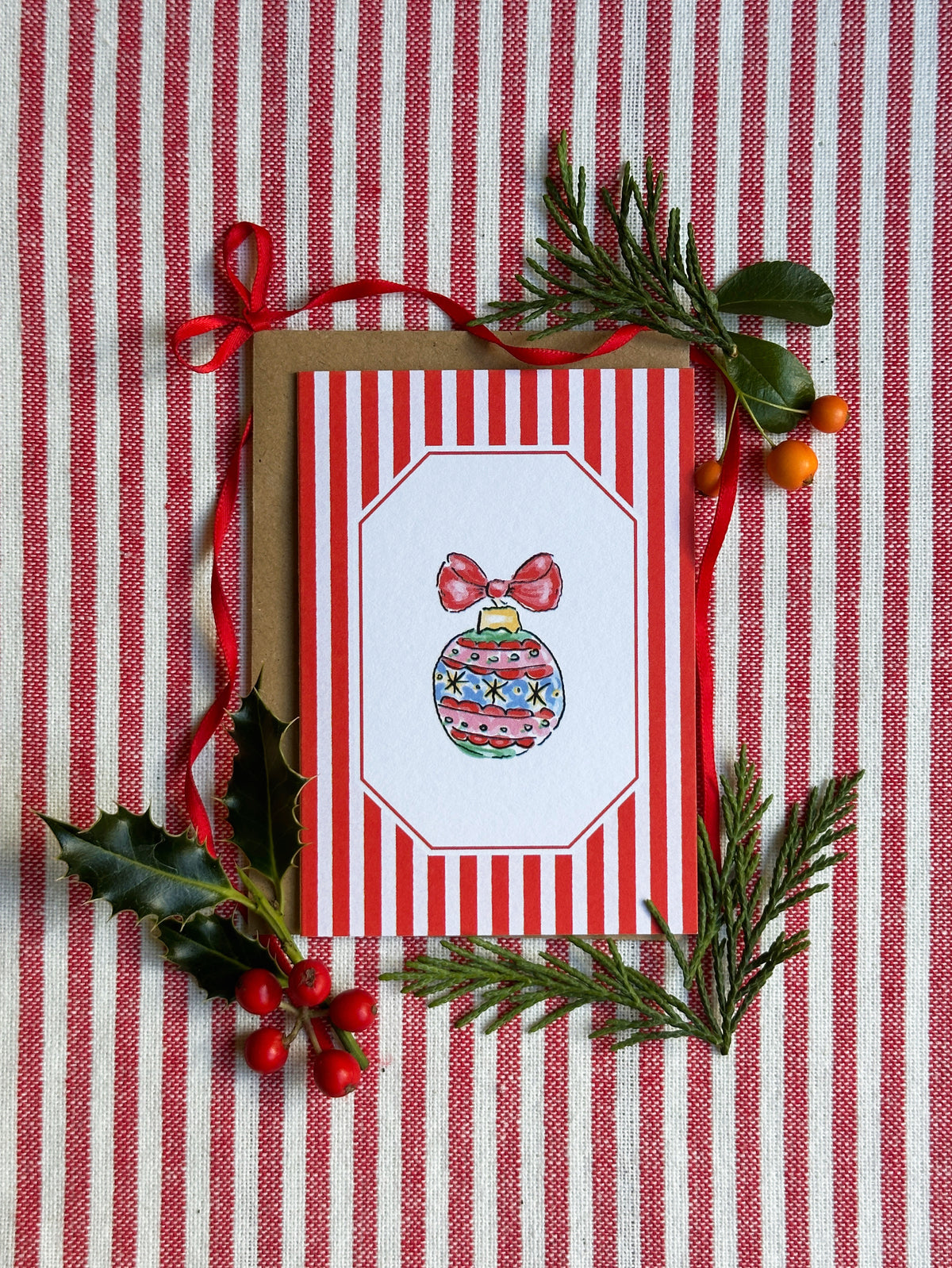 A Christmas greeting card with red and white vertical stripes and an illustration of a decorated ornament, styled with a kraft envelope, red ribbon, holly leaves, pine branches, and berries on a red-and-white striped fabric background.