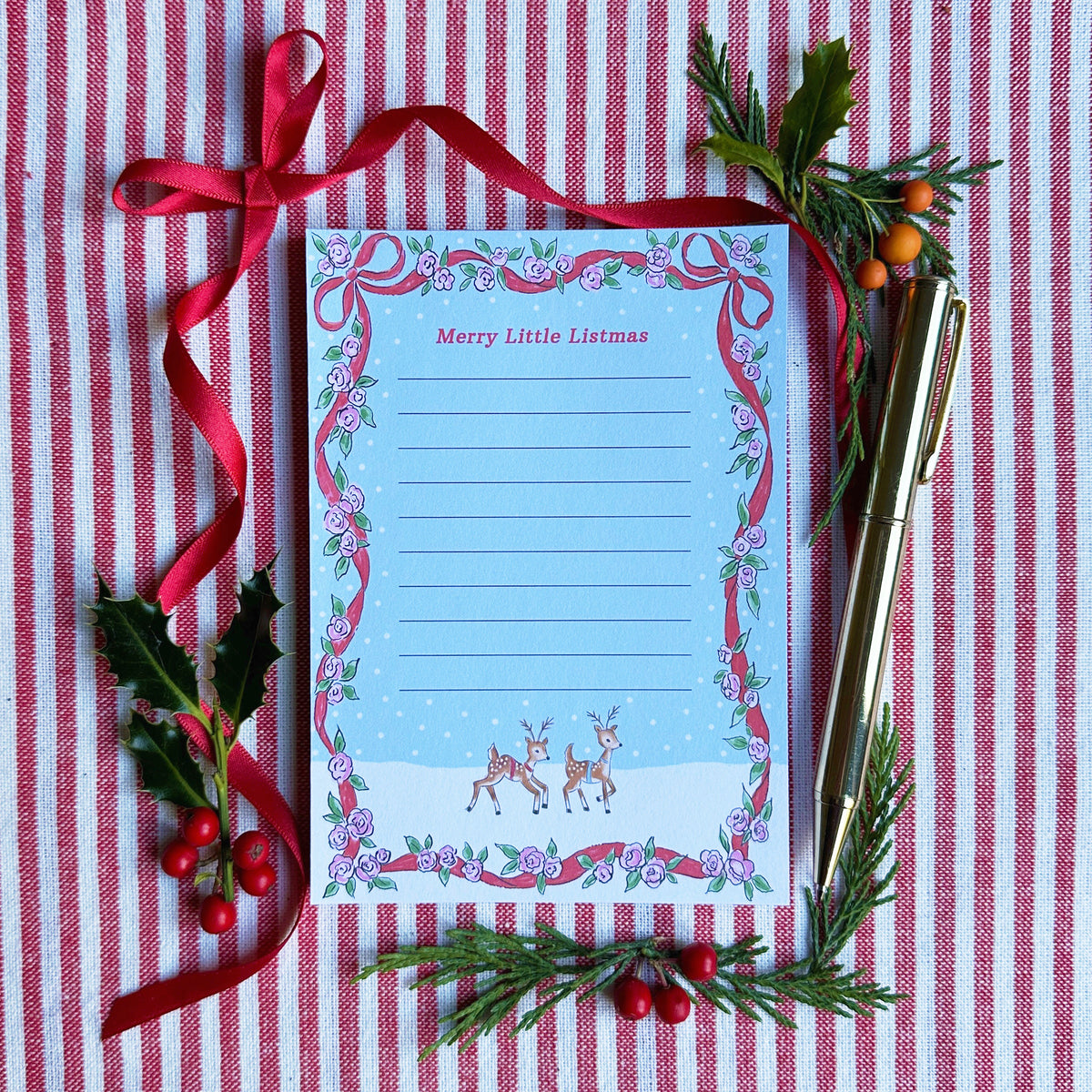 A6 Tear Off sheet Notepad with red ribbon and floral border blue background and two deer. Top of Notepad reads 'Merry Little Listmas". Backdrop is styled on a red and white striped fabric, gold pen, red ribbon and greenery and berries.