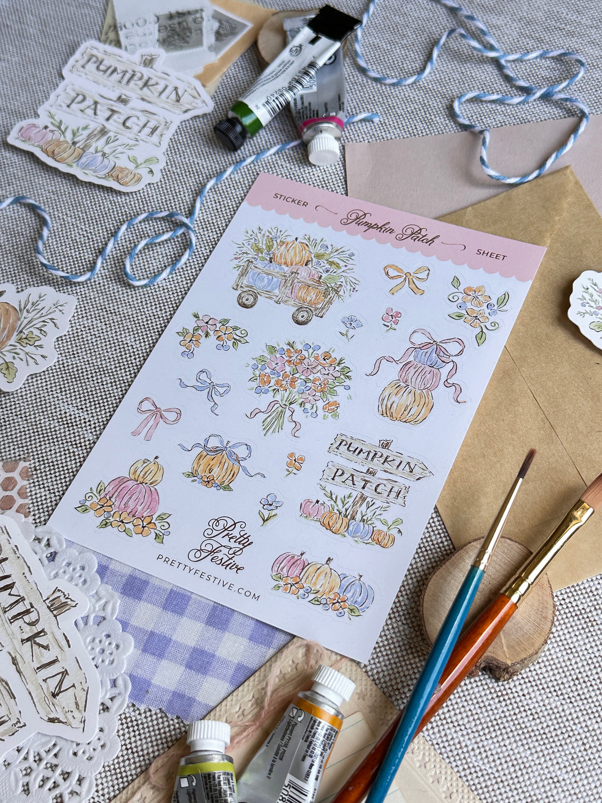 A sheet of decorative stickers featuring pumpkins, flowers, bows, and the words 'Pumpkin Patch' arranged on a styled craft surface with paintbrushes, paint tubes, envelopes, and ribbon.