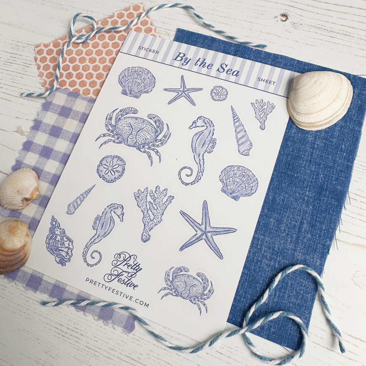 A sheet of decorative stickers featuring nautical designs like crabs, starfish, seahorses, shells, and coral in blue line art, styled on a white wood surface with blue-and-white twine, a gingham cloth, patterned paper, and seashells.