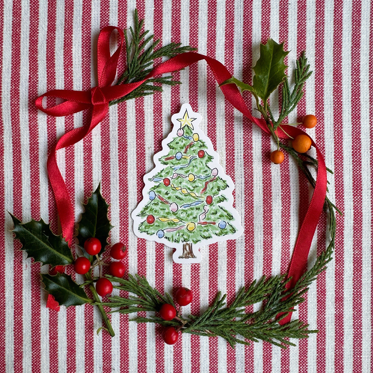 Vinyl die cut sticker of illustrated christmas tree. Background is red and white striped fabric with red ribbon greenery and berries.