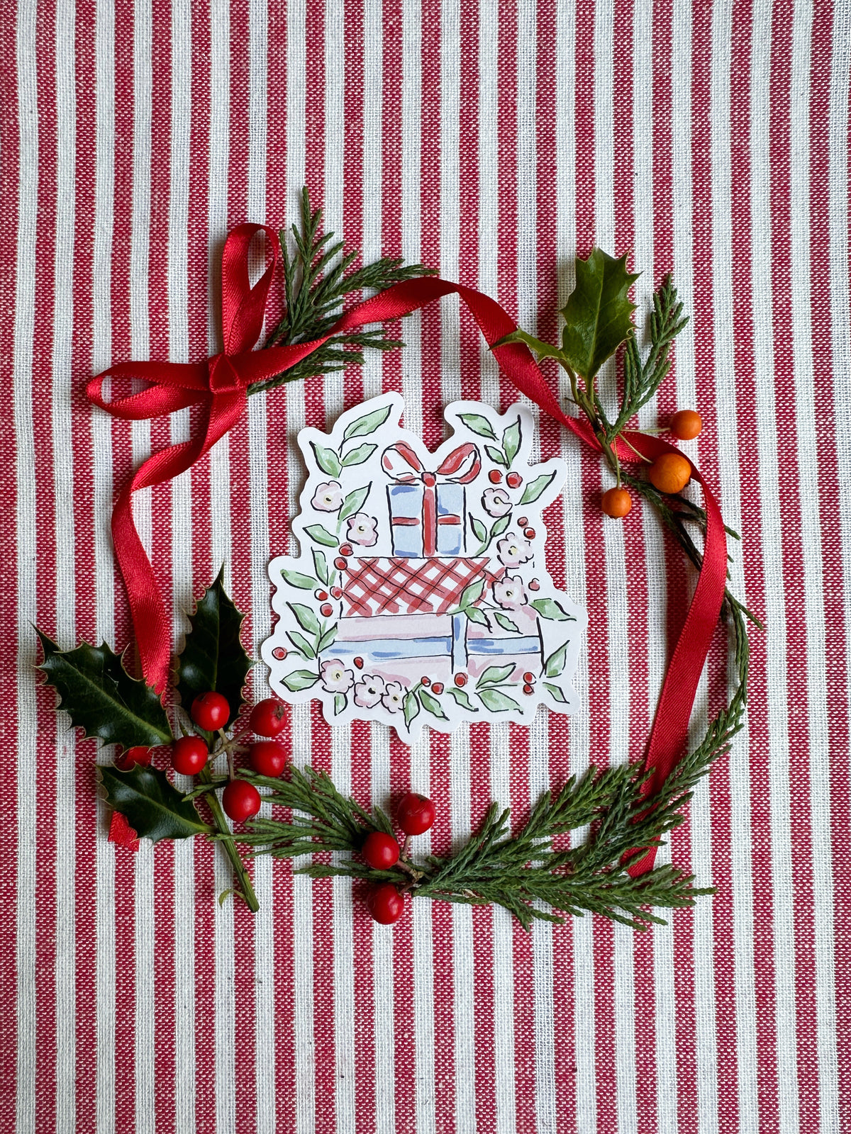 Vinyl die cut sticker of illustrated christmas gift stack surrounded by foliage and flowers. The background is red and white striped fabric with red ribbon greenery and berries.