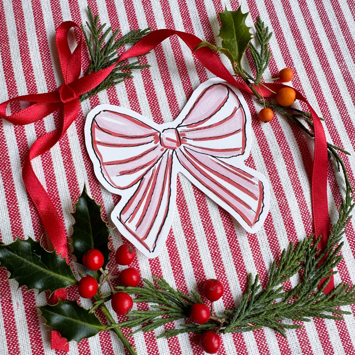 Vinyl die cut sticker of illustrated pink red and white bow. Background is red and white striped fabric with red ribbon greenery and berries.