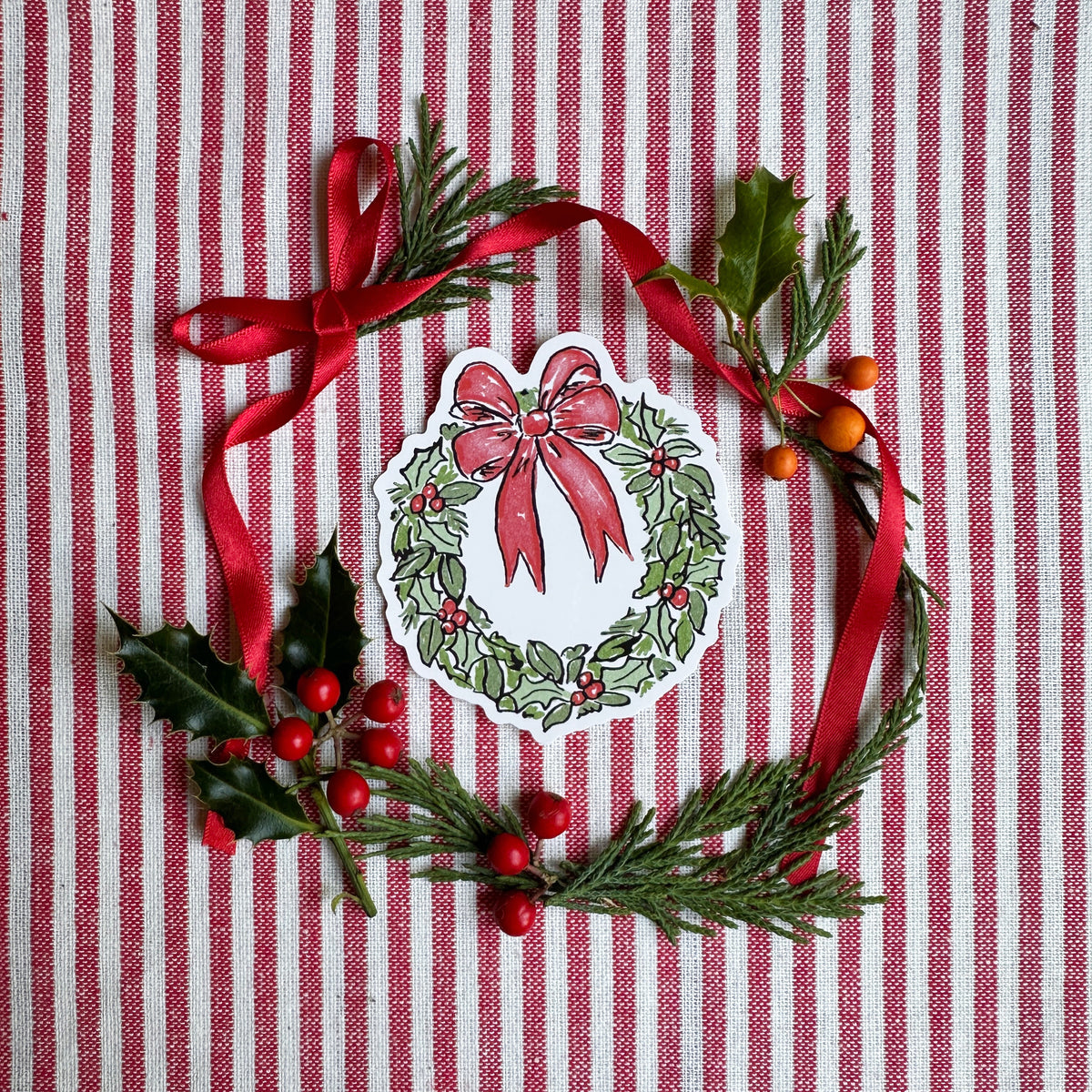 Vinyl die cut sticker of illustrated christmas wreath with red bow. Background is red and white striped fabric with red ribbon greenery and berries.