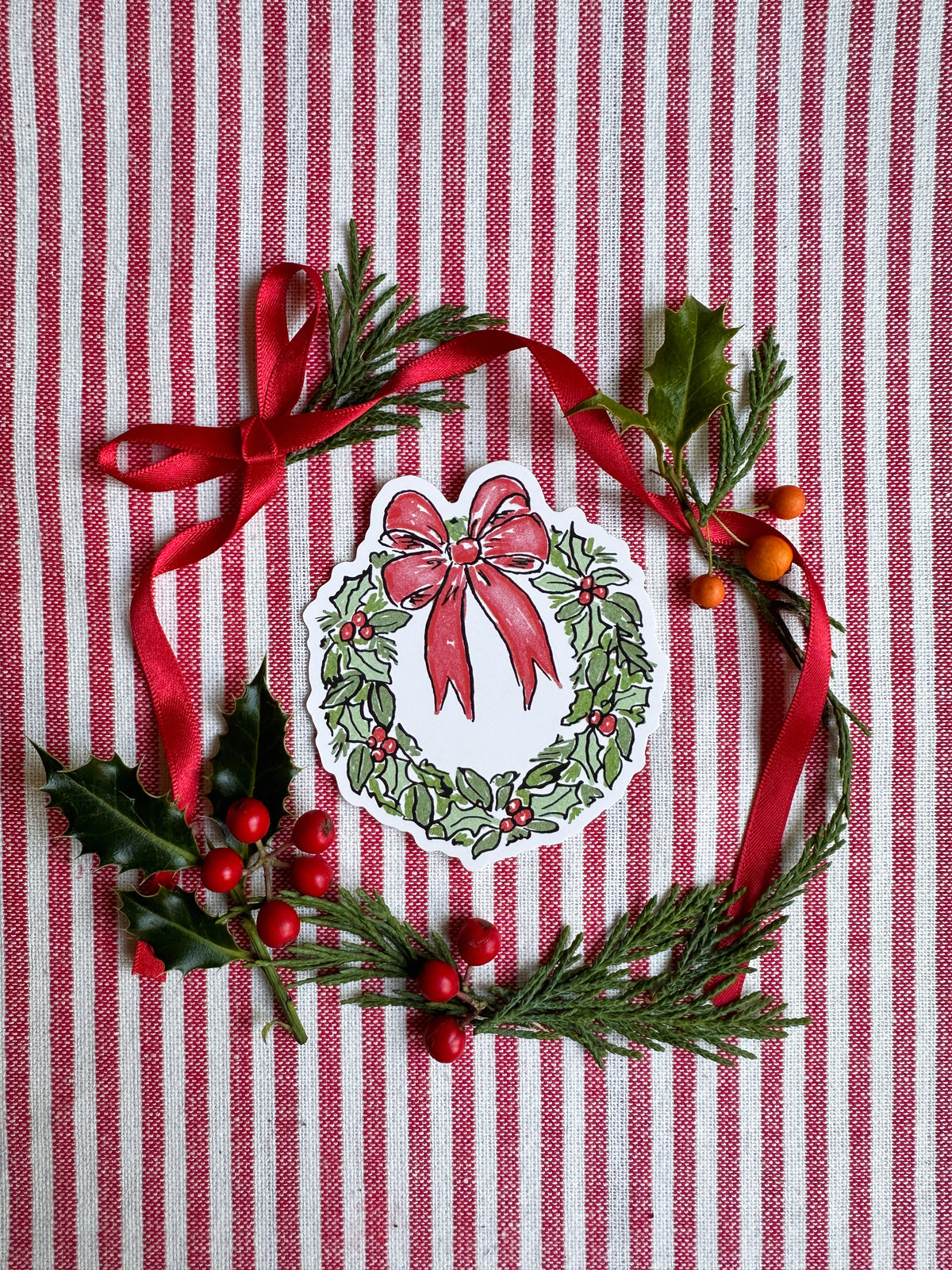 Vinyl die cut sticker of illustrated christmas wreath with red bow. Background is red and white striped fabric with red ribbon greenery and berries.