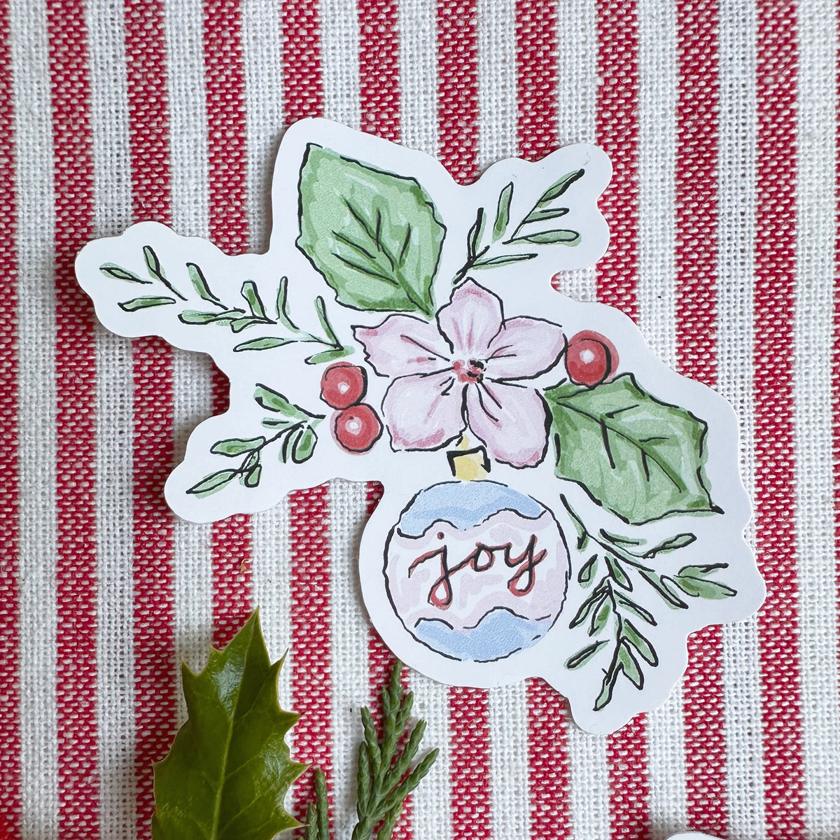 Vinyl die cut sticker of illustrated christmas ornament with foliage, berries and flowers. The background is red and white striped fabric with red ribbon greenery and berries.