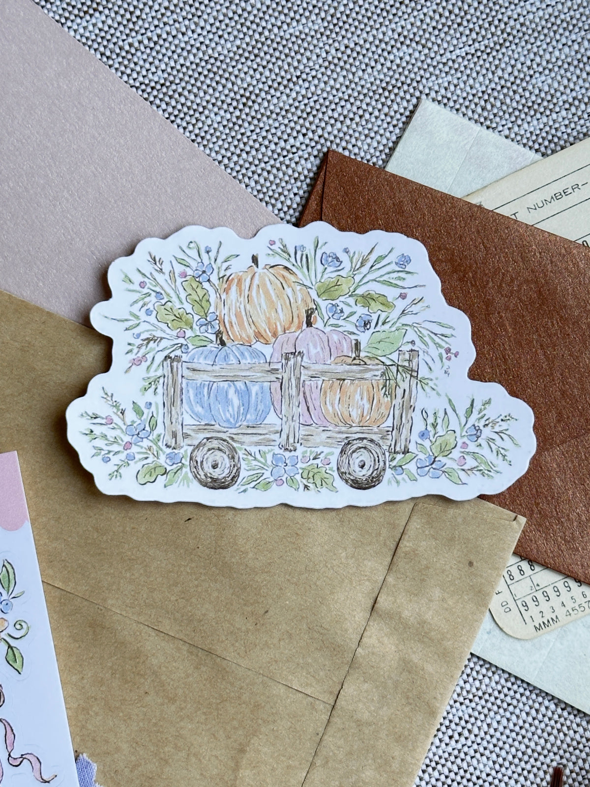 A scalloped die-cut sticker featuring an autumn watercolor illustration of pumpkins in a wooden wagon surrounded by leaves and flowers, displayed on brown kraft paper envelopes over a textured gray fabric background.
