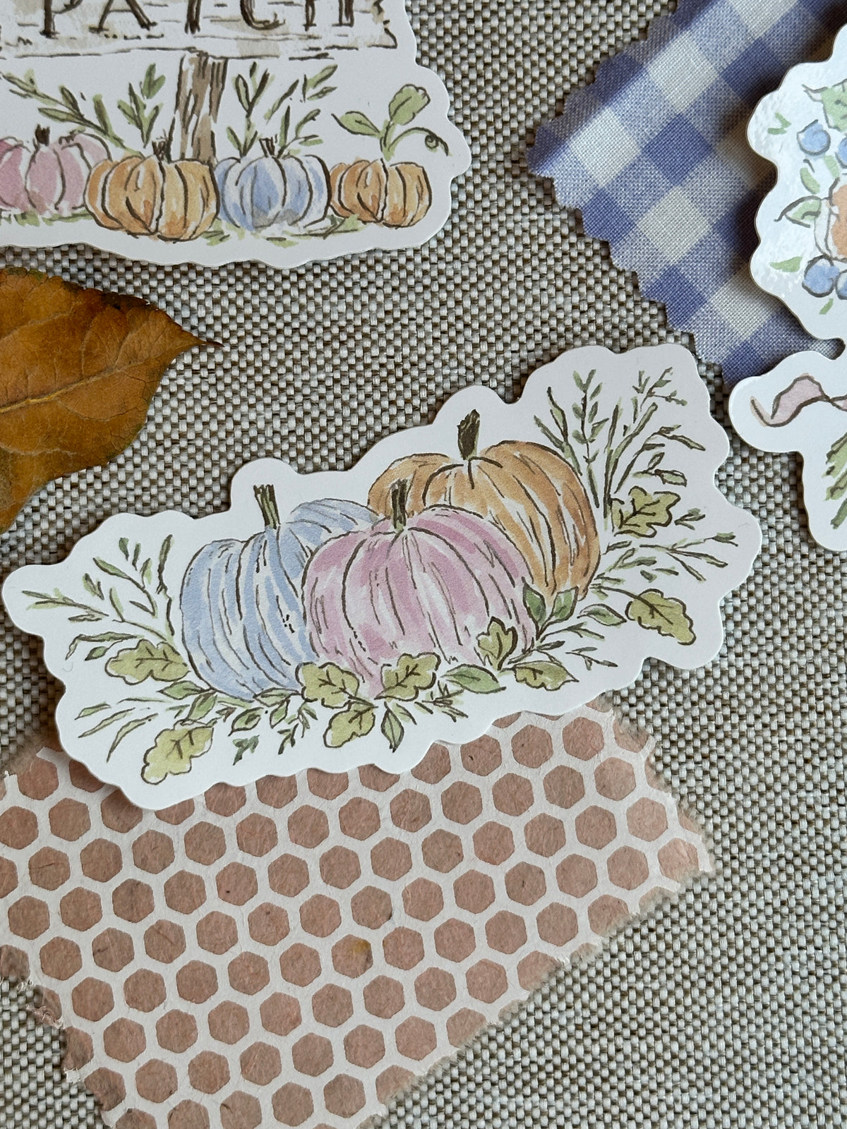 A close-up of a die-cut sticker featuring three hand-painted style fall pumpkins in pastel blue, pink, and orange with green leaves, placed on a textured beige fabric background with other autumn-themed paper pieces nearby.