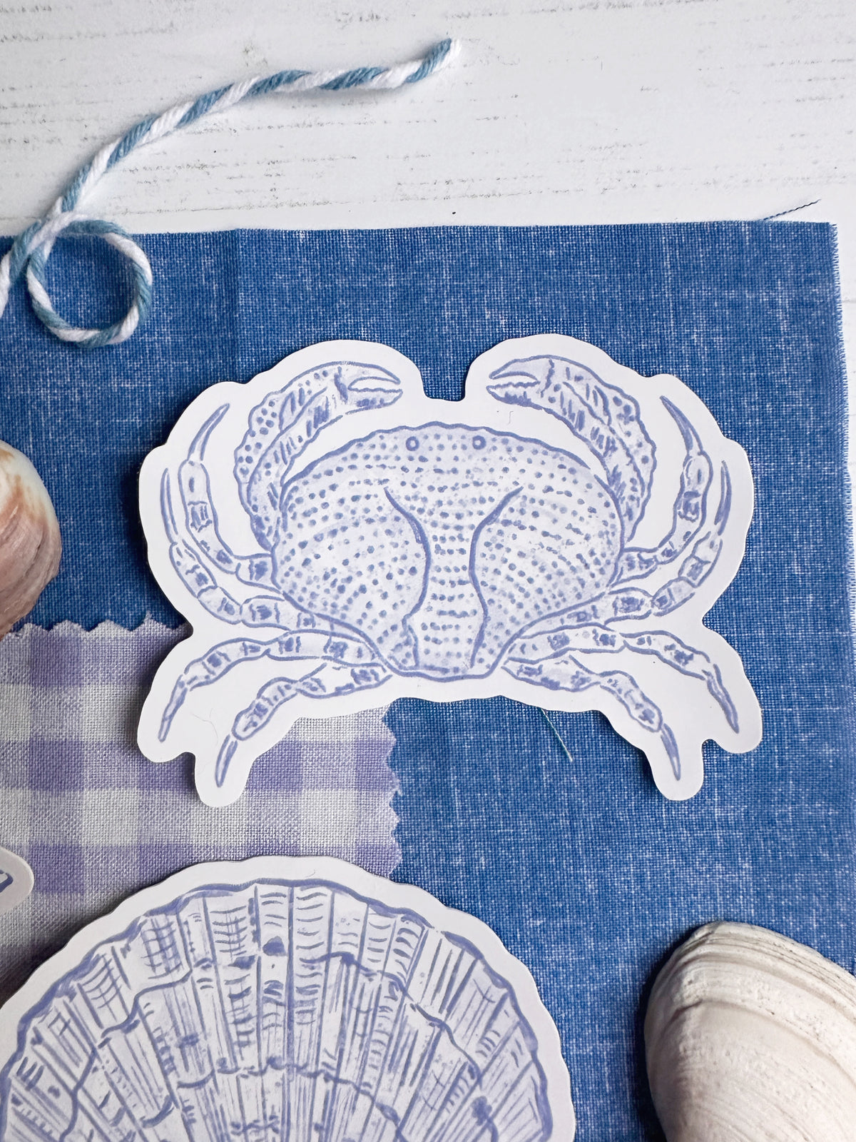 A die-cut sticker featuring a blue and white patterned crab, displayed on a blue linen background with a matching blue-and-white gingham cloth, a shell, and a piece of string.