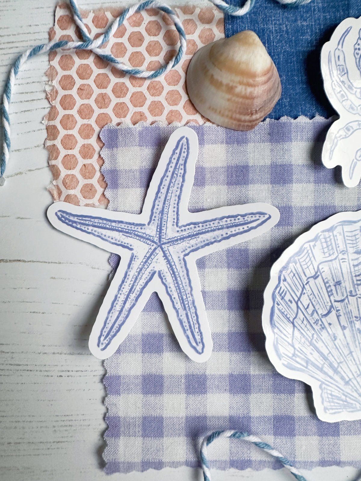 A styled flat-lay featuring die-cut stickers shaped like a blue starfish and a white seashell with blue line art, arranged on a lavender and white gingham fabric over a peach honeycomb patterned cloth, with a real seashell prop and blue string.