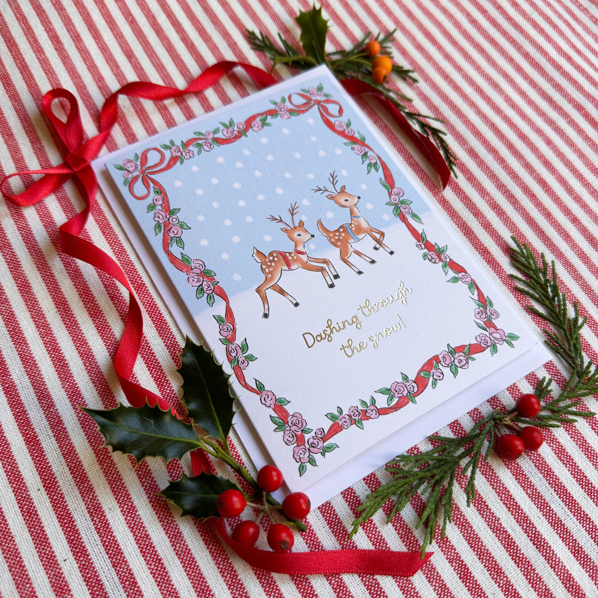 christmas card of two reindeer caption Dashing Through the Snow. surrounded by bows and evergreens on a red and white striped backdrop