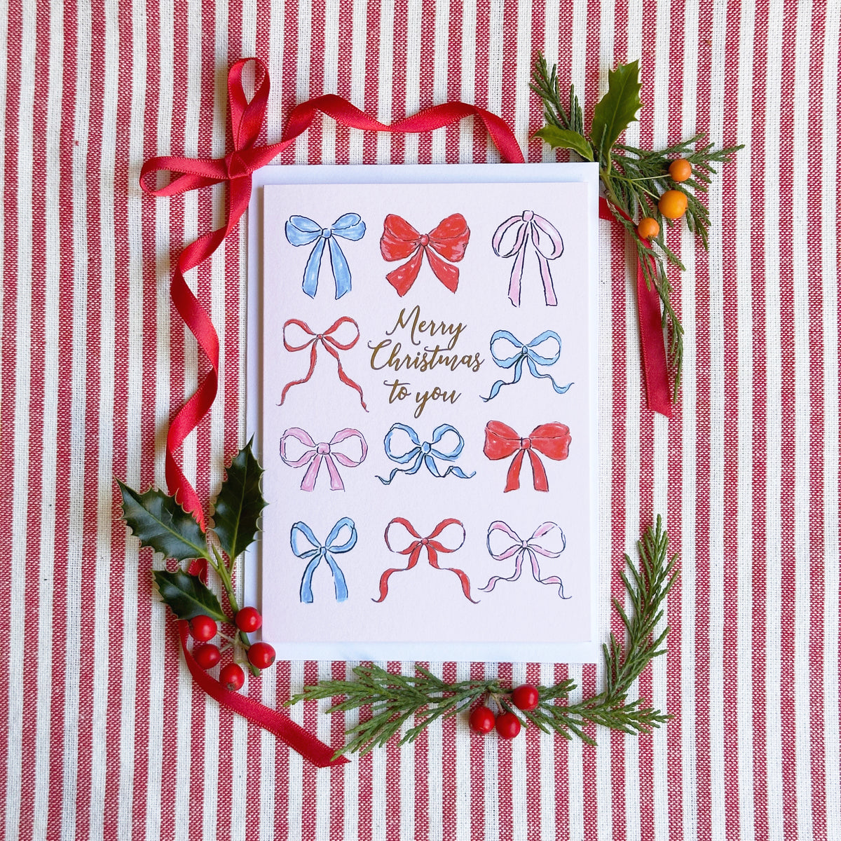 a christmas card with a soft pink background with several illustrated bows and captioned Merry Christmas to You. Background of image is red ribbon, foliage and berries on a red and white striped backgroumd