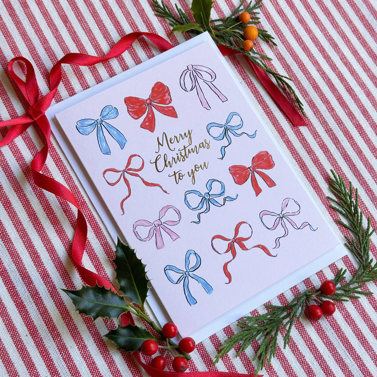 a christmas card with a soft pink background with several illustrated bows and captioned Merry Christmas to You. Background of image is red ribbon, foliage and berries on a red and white striped backgroumd
