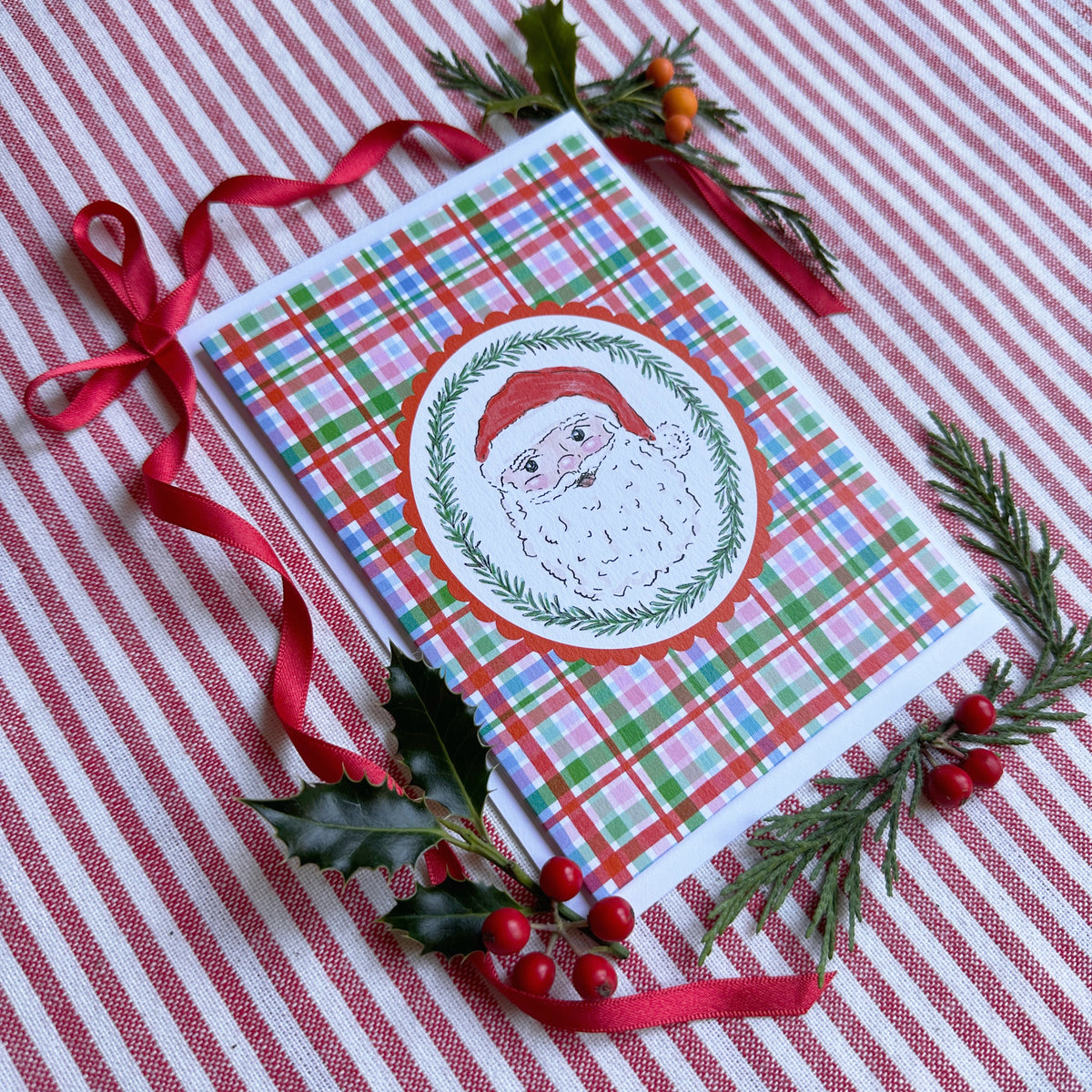 christmas card of Santa head surrounded by bows and evergreens on a red and white striped backdrop