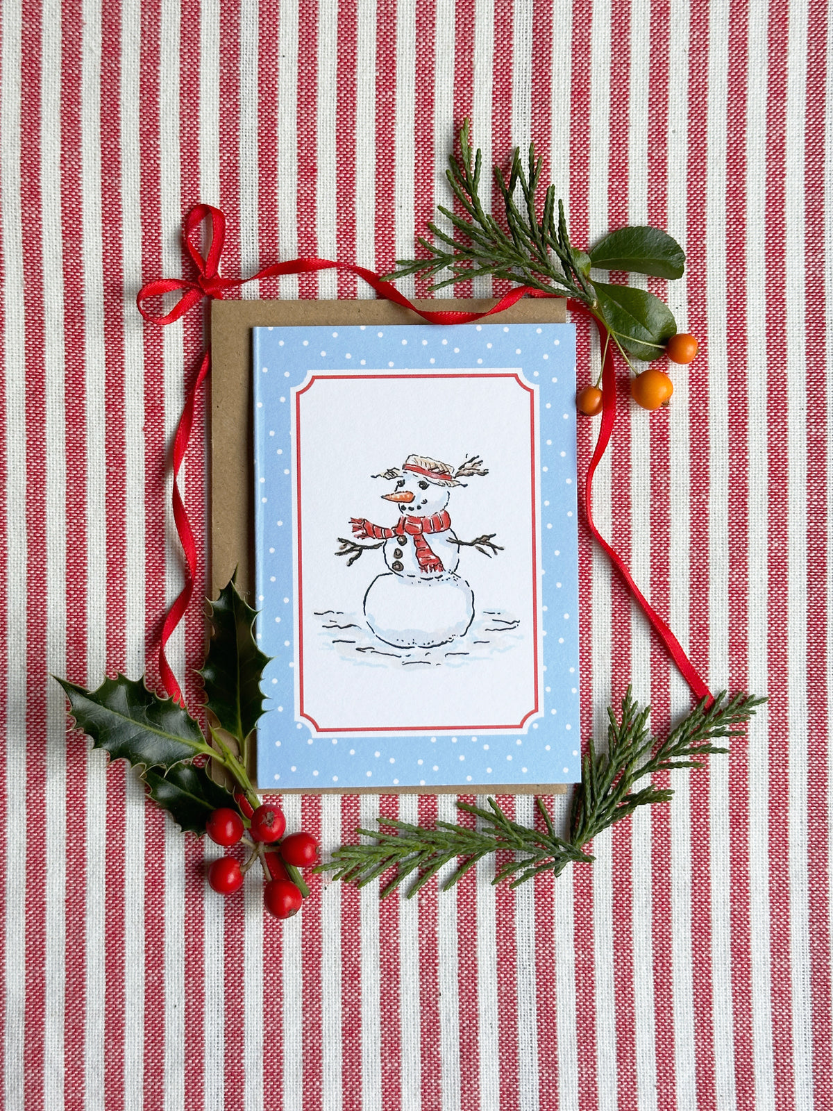 A mini Christmas greeting card with a snowman illustration on a light blue background, styled on a red and white striped fabric with pine branches, holly leaves, and red ribbon.