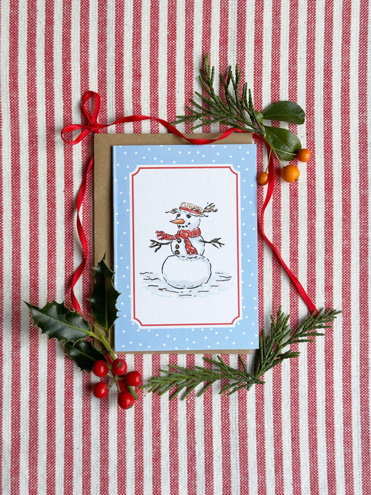 A mini Christmas greeting card with a snowman illustration on a light blue background, styled on a red and white striped fabric with pine branches, holly leaves, and red ribbon.
