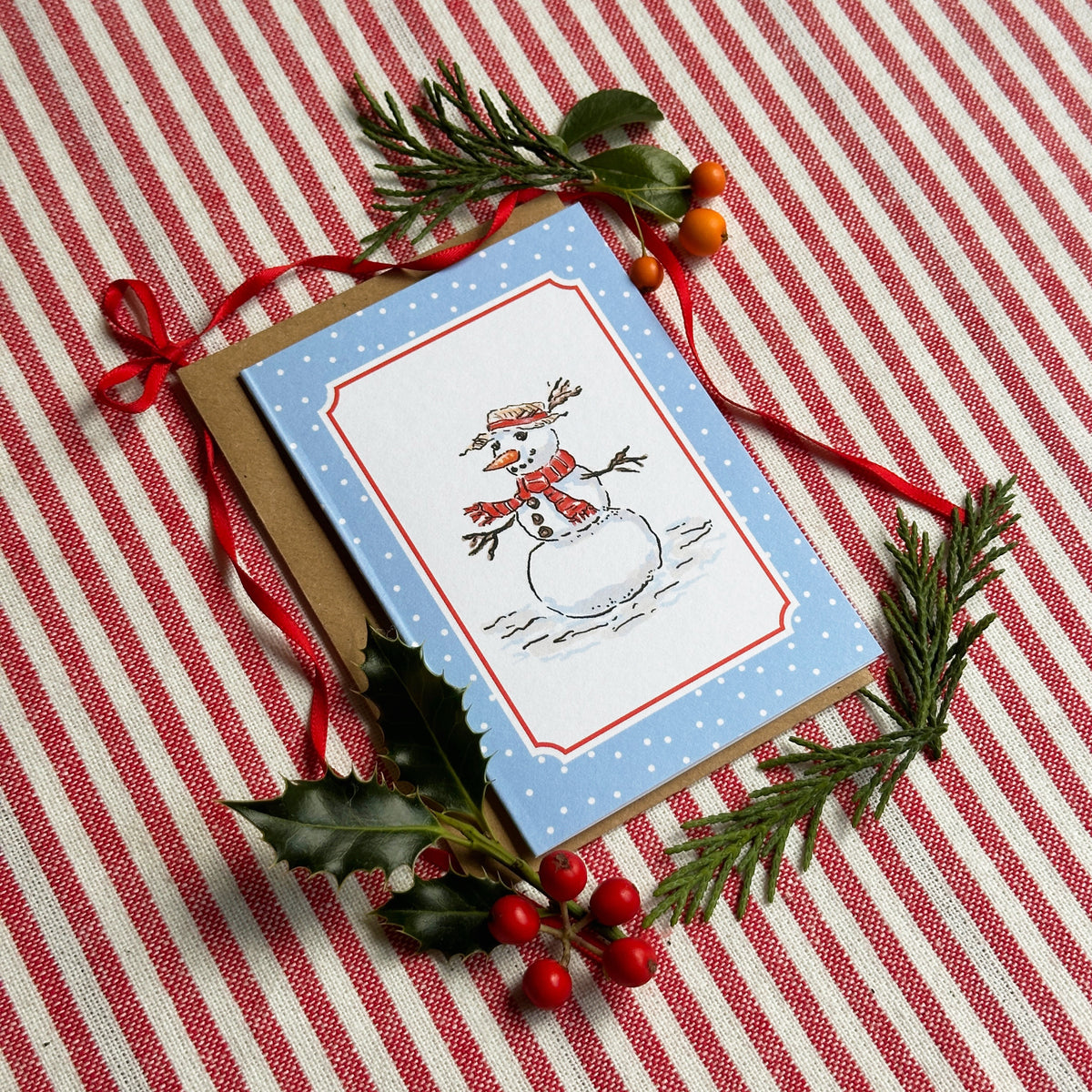 A mini Christmas greeting card with a snowman illustration on a light blue background, styled on a red and white striped fabric with pine branches, holly leaves, and red ribbon.