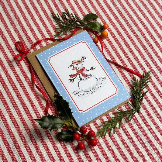 A mini Christmas greeting card with a snowman illustration on a light blue background, styled on a red and white striped fabric with pine branches, holly leaves, and red ribbon.