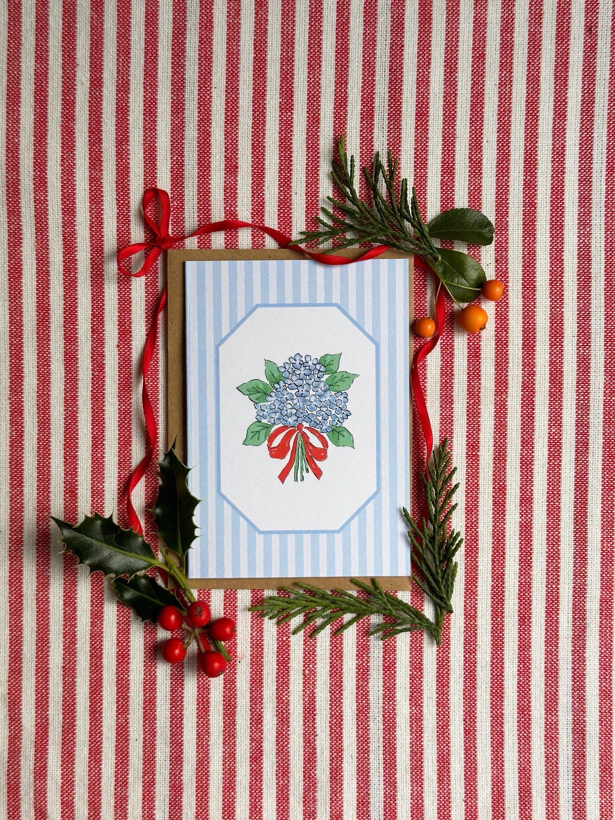 A square greeting card with a blue and white striped border and a central illustration of a blue hydrangea with green leaves and a red ribbon, styled on a red and white striped fabric background with holly, pine branches, and red berries.
