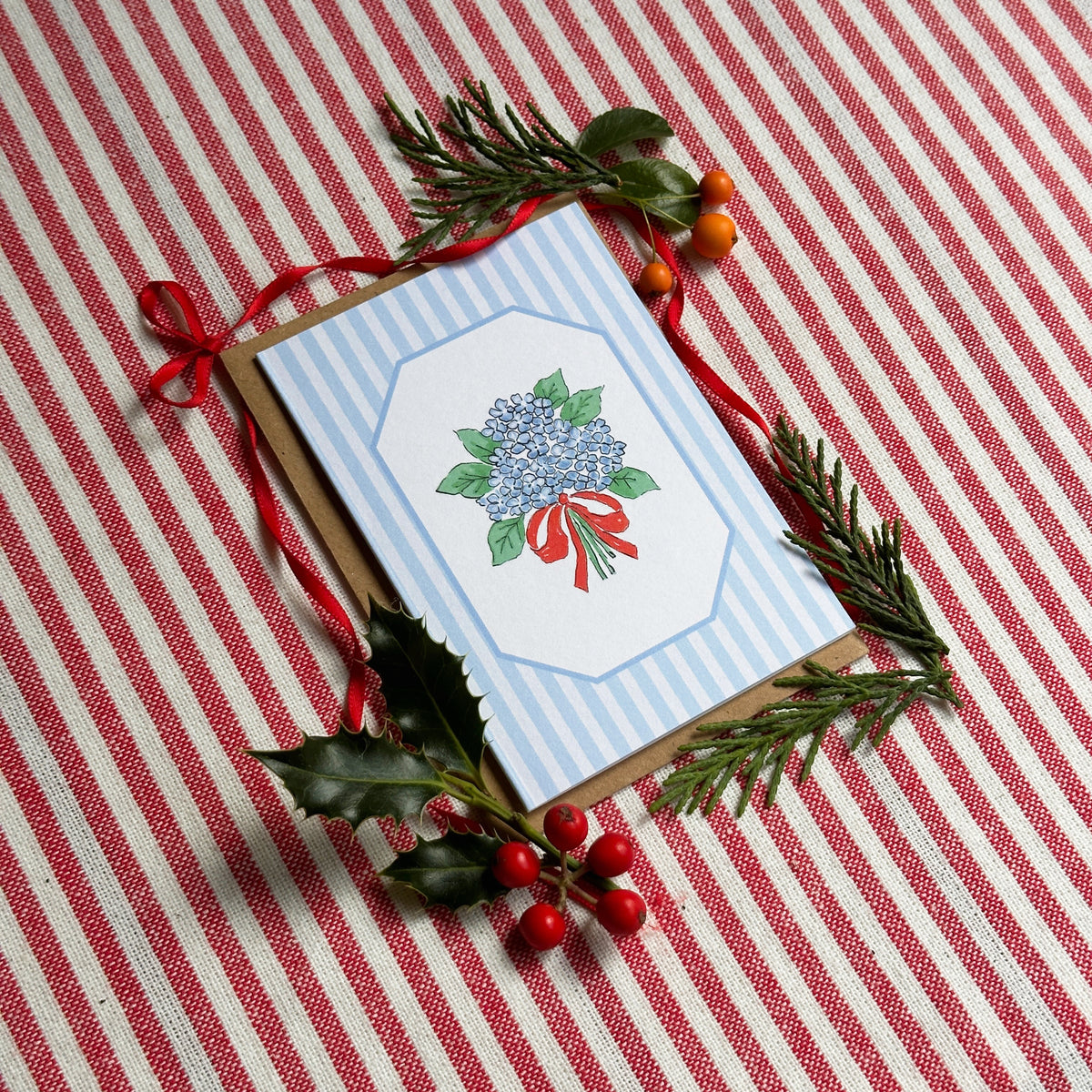 A mini greeting card with a blue and white striped border and an illustration of a blue hydrangea with a red ribbon, styled on a red and white striped tablecloth with pine branches, holly leaves, and red berries.