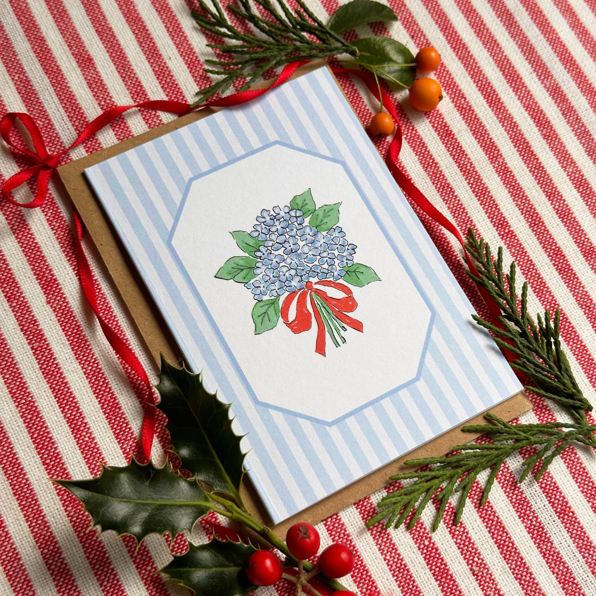 A mini greeting card with a blue and white striped border and an illustration of a blue hydrangea bouquet with green leaves and a red ribbon, placed on a red and white striped fabric with holly and pine branches around it.