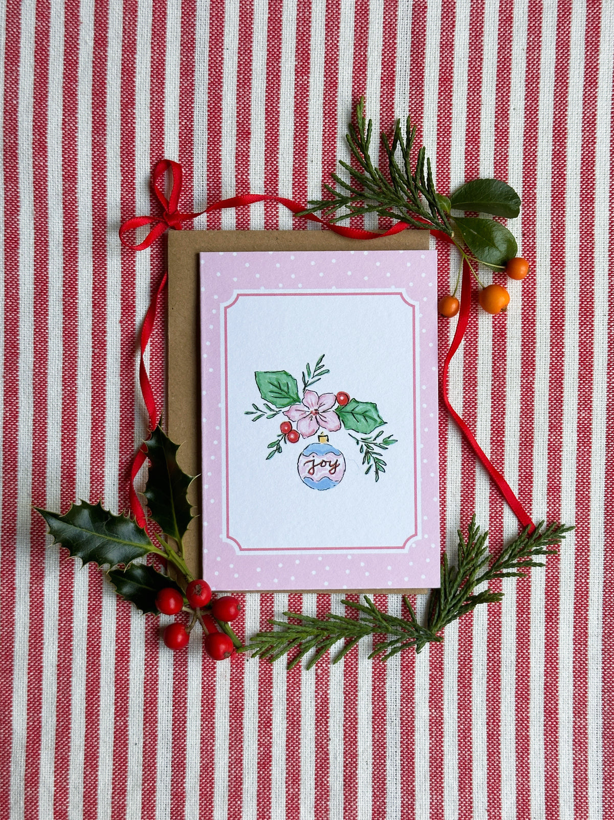 A mini greeting card with a pink border and a small Christmas illustration, placed on a red-and-white striped fabric background and styled with a kraft envelope, red ribbon, holly leaves, pine branches, and small orange berries.