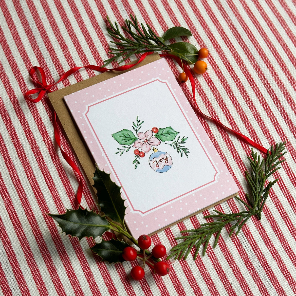 A mini A7 Christmas greeting card with a pink border and a central illustration of a decorated ornament, placed on a brown envelope over a red-and-white striped fabric background with holly and pine sprigs.
