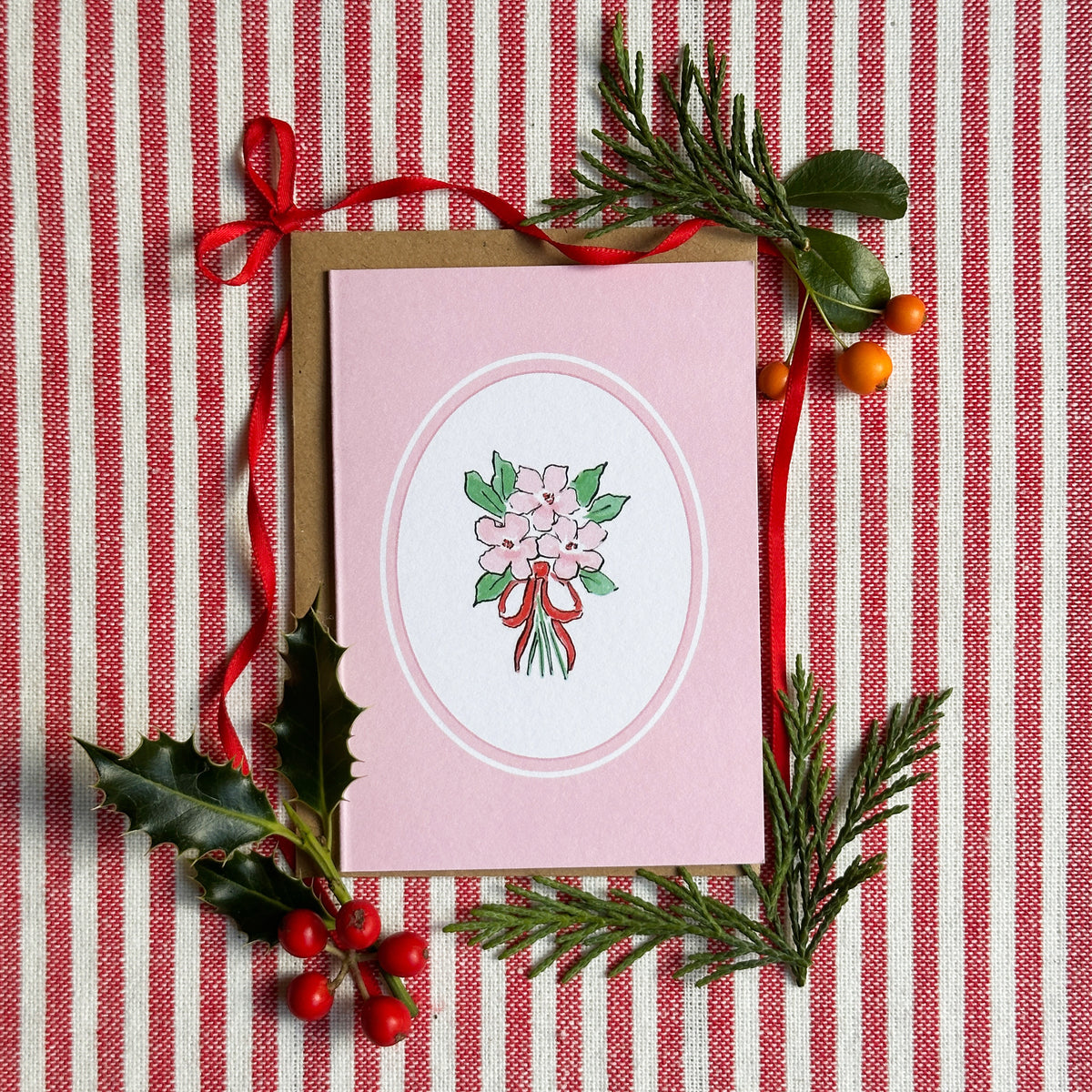 A pink greeting card with a white oval center featuring a bouquet illustration, styled on a red-and-white striped fabric with holly, pine branches, and red ribbon.