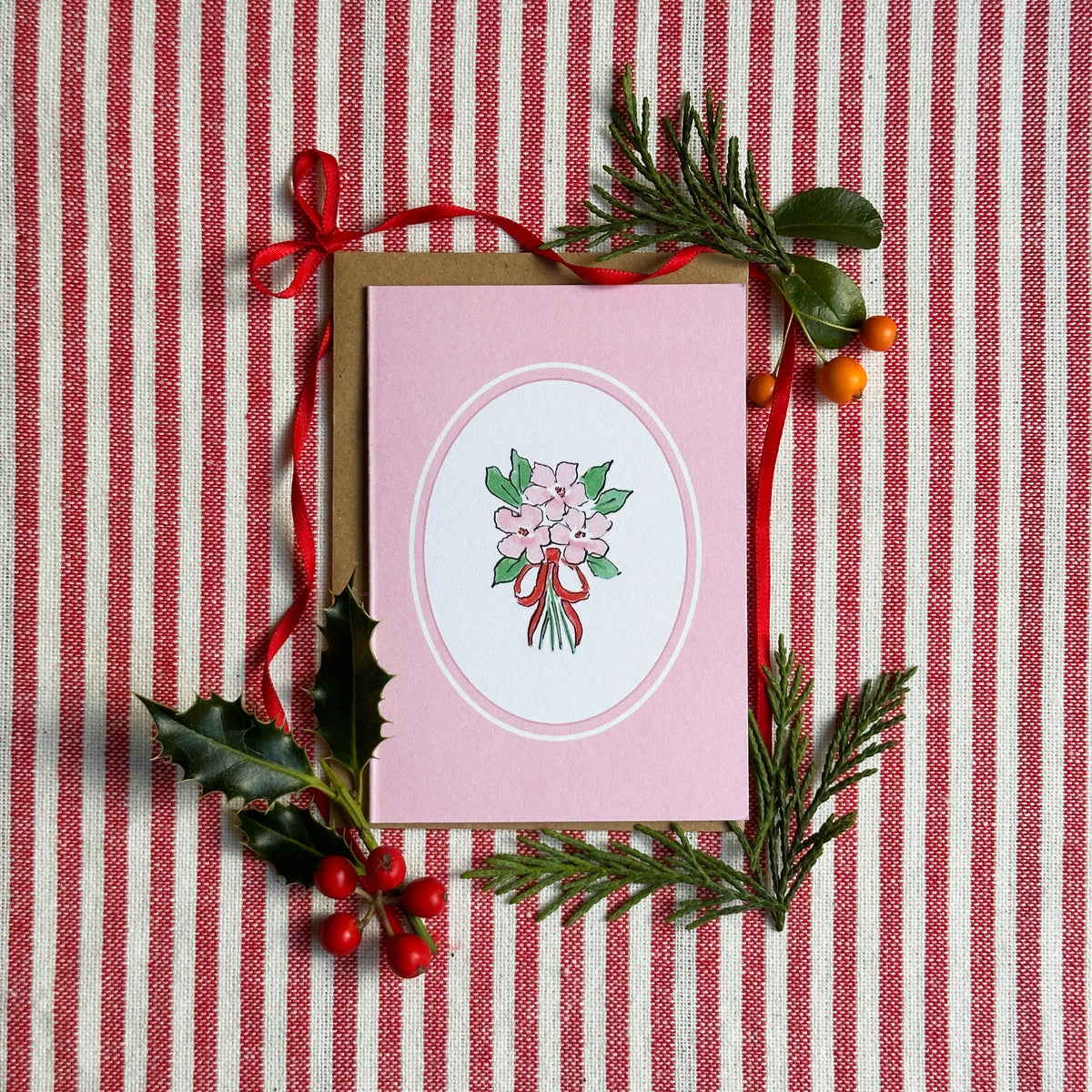 A pink greeting card with a white oval center featuring a bouquet illustration, styled on a red and white striped fabric background with a brown envelope, red ribbon, and green holly and pine branches.
