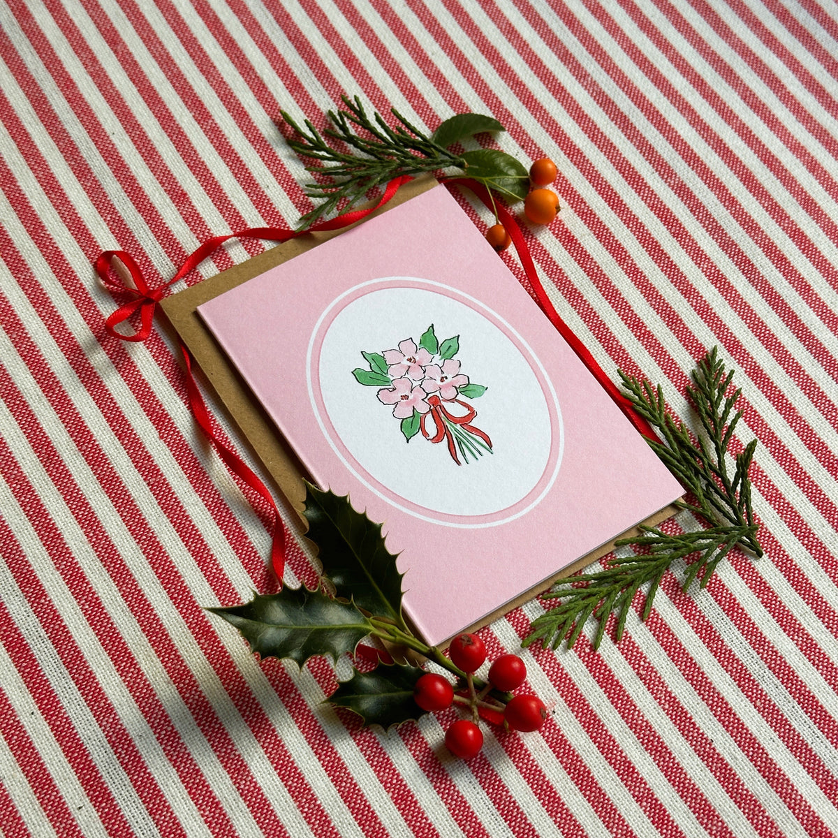 A pink greeting card with a circular floral illustration in the center, placed on a red-and-white striped fabric background and styled with green pine branches, holly leaves, and red berries.