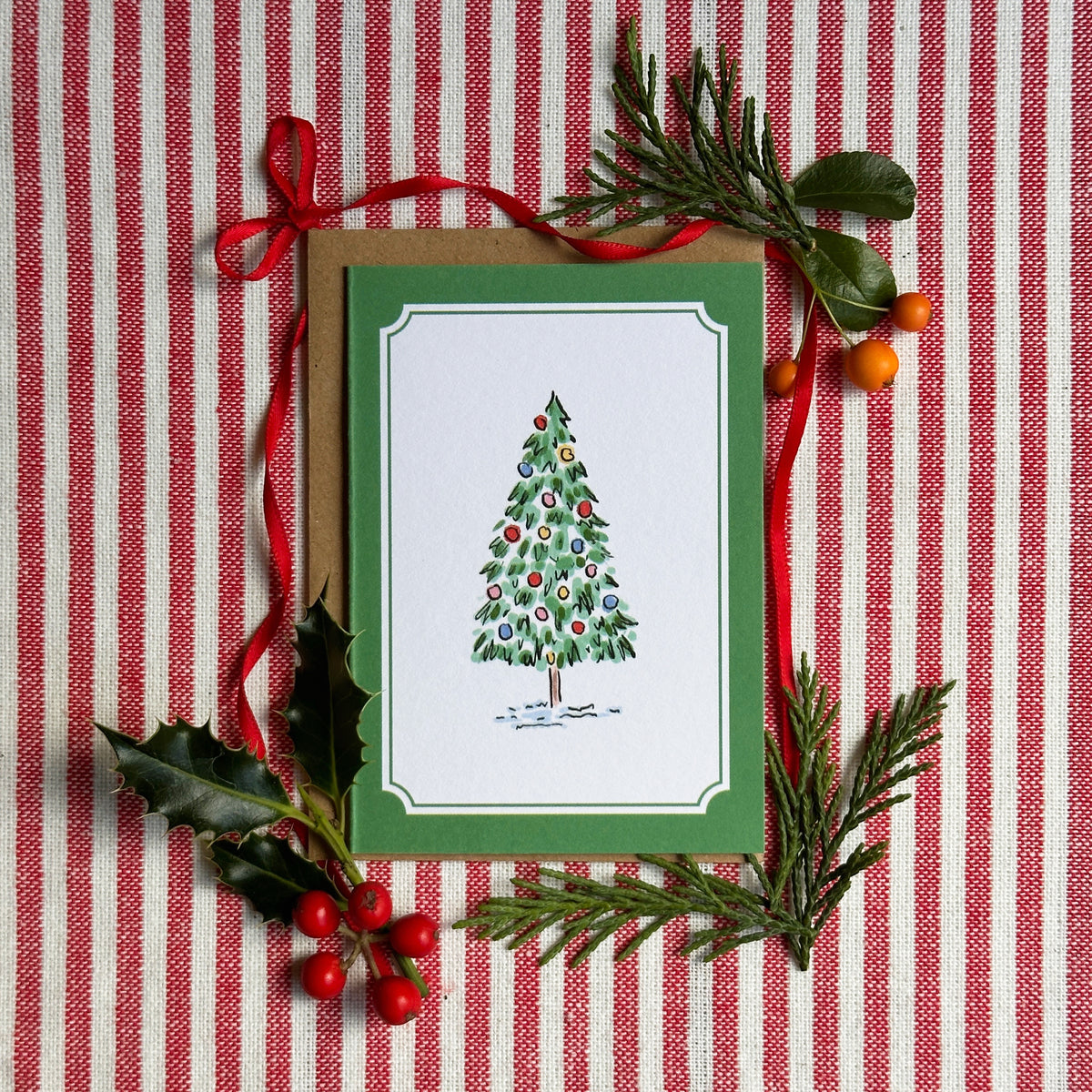 A mini Christmas greeting card with a green border and an illustration of a decorated tree, styled on a red-and-white striped fabric with pine branches, holly leaves, and red ribbon.