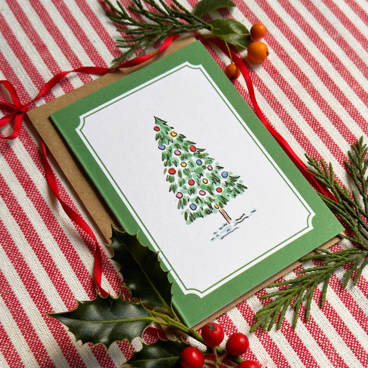 A mini Christmas greeting card with a green border and an illustration of a decorated tree, styled on a red-and-white striped fabric with pine branches, holly leaves, and red ribbon.