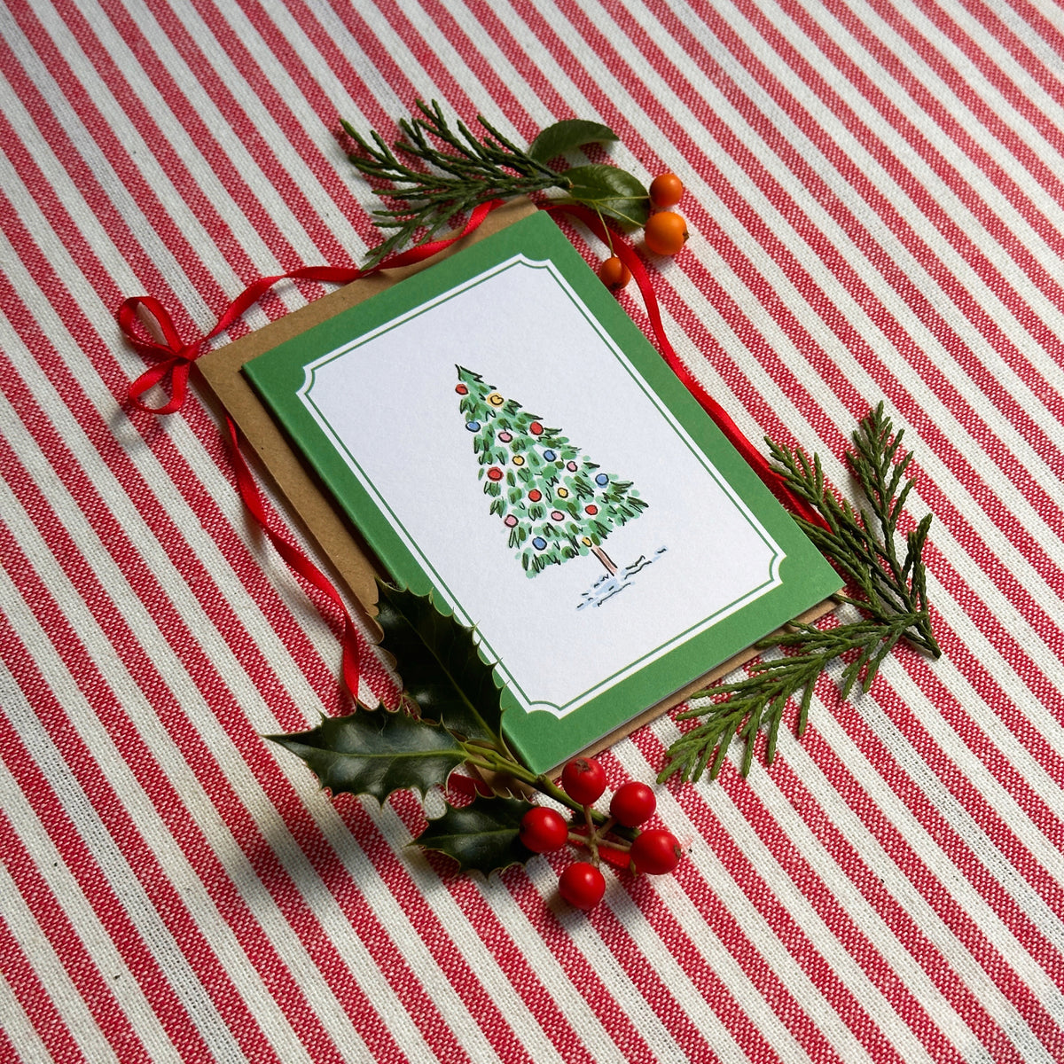 A Christmas greeting card with a green border and an illustration of a decorated Christmas tree, styled on a red and white striped fabric background with pine branches, holly leaves, and red berries.