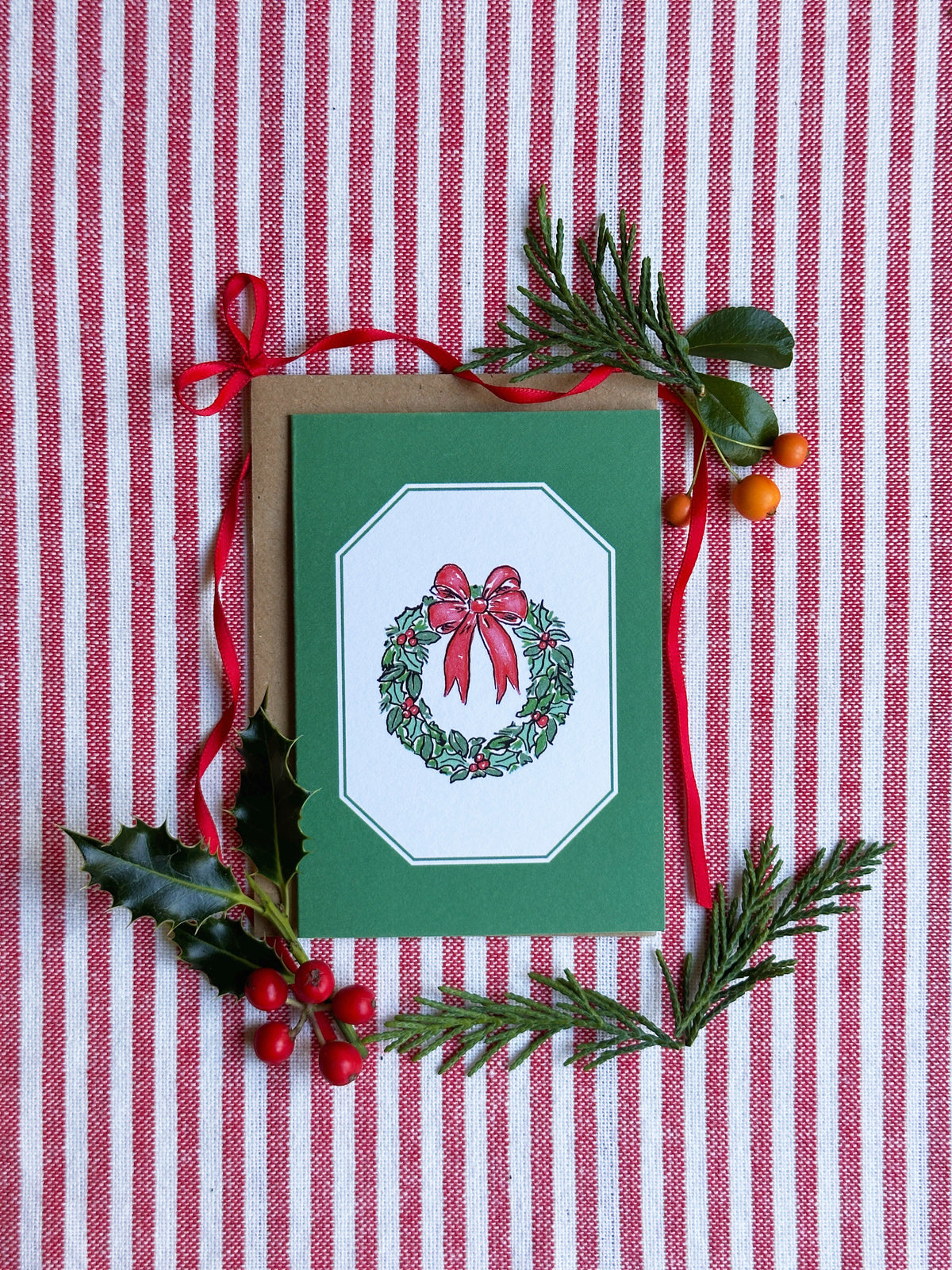 A green Christmas greeting card with a wreath and red bow illustration, styled on a red and white striped fabric with pine branches, holly leaves, and red berries.