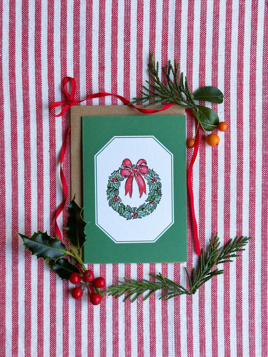 A green Christmas greeting card with a wreath and red bow illustration, styled on a red and white striped fabric with pine branches, holly leaves, and red berries.