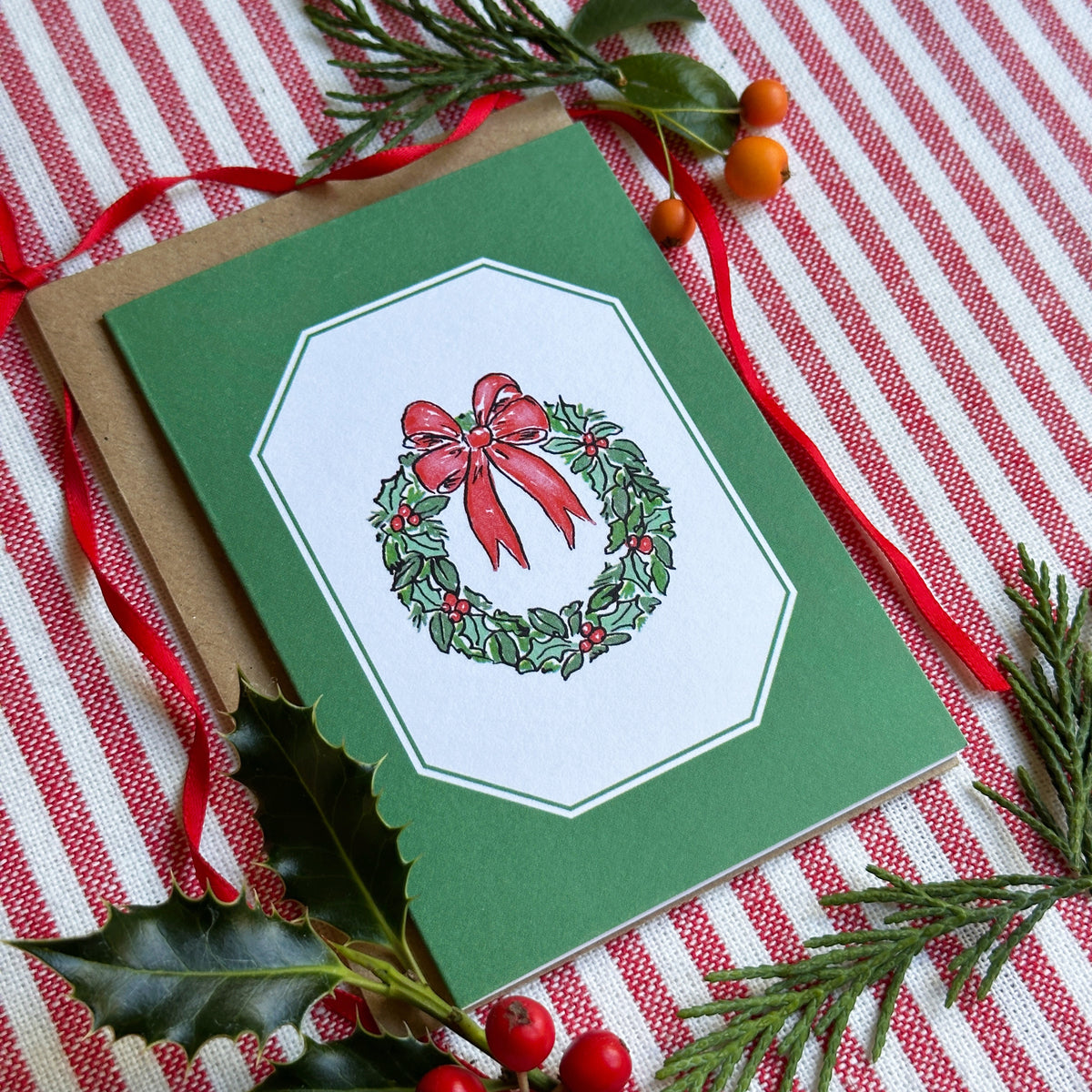 A green Christmas greeting card with a white octagonal center panel featuring a red bow and holly wreath illustration, placed on a kraft envelope over a red-and-white striped fabric with pine branches and holly leaves.