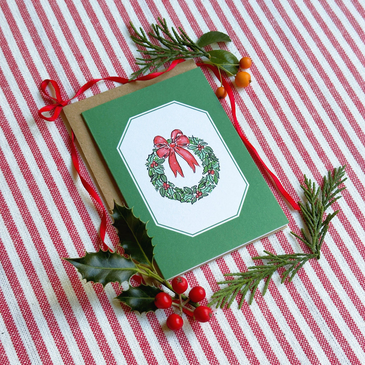 A green Christmas greeting card with a wreath illustration, placed on a red-and-white striped fabric background with pine branches, holly leaves, and red berries as styling props.