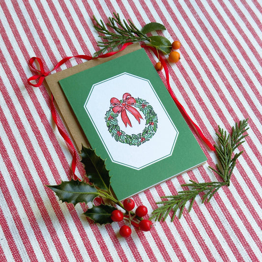 A green Christmas greeting card with a wreath illustration, placed on a red-and-white striped fabric background with pine branches, holly leaves, and red berries as styling props.