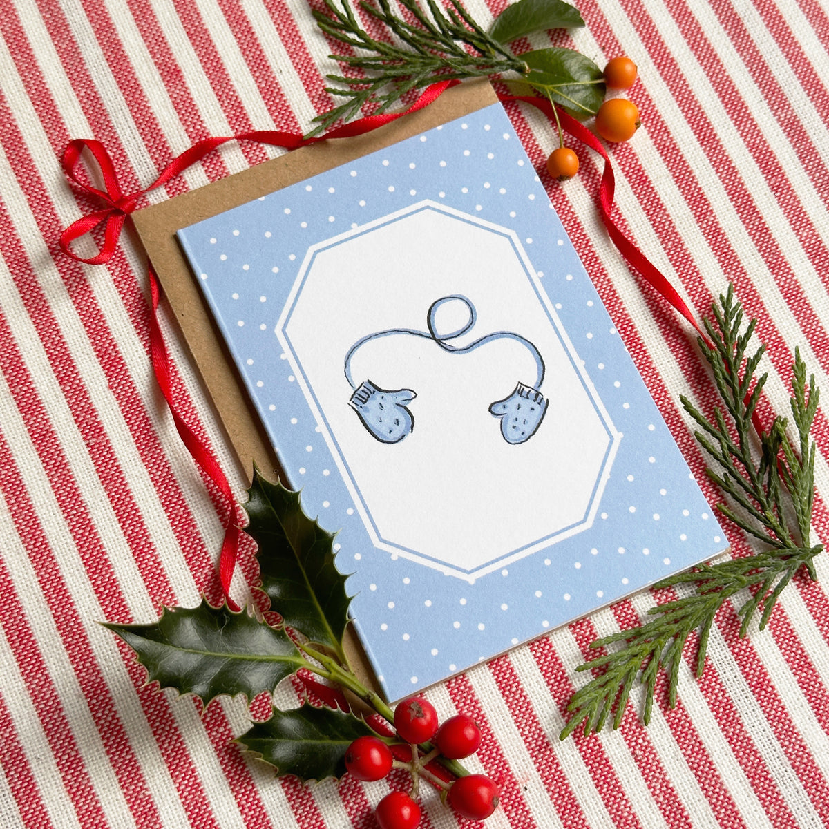A mini A7 greeting card with a light blue and white polka-dot border and a central white octagonal panel showing a blue mitten illustration. The card is placed on a red-and-white striped fabric and surrounded by holiday greenery, red berries, and red ribbon.